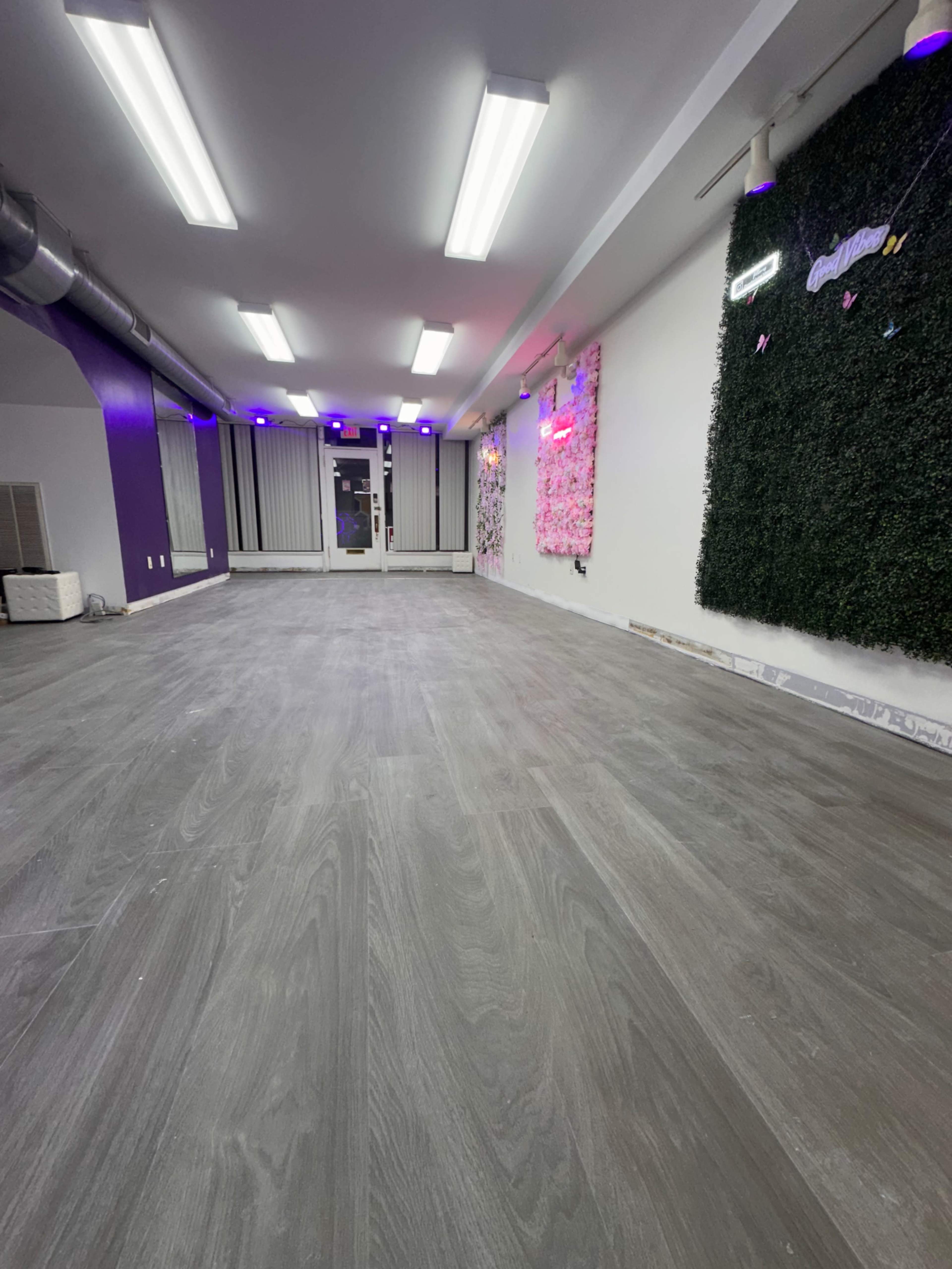 The image shows a spacious, vacant room with a grey wooden floor, bright ceiling lights, and a wall featuring greenery and neon signs.