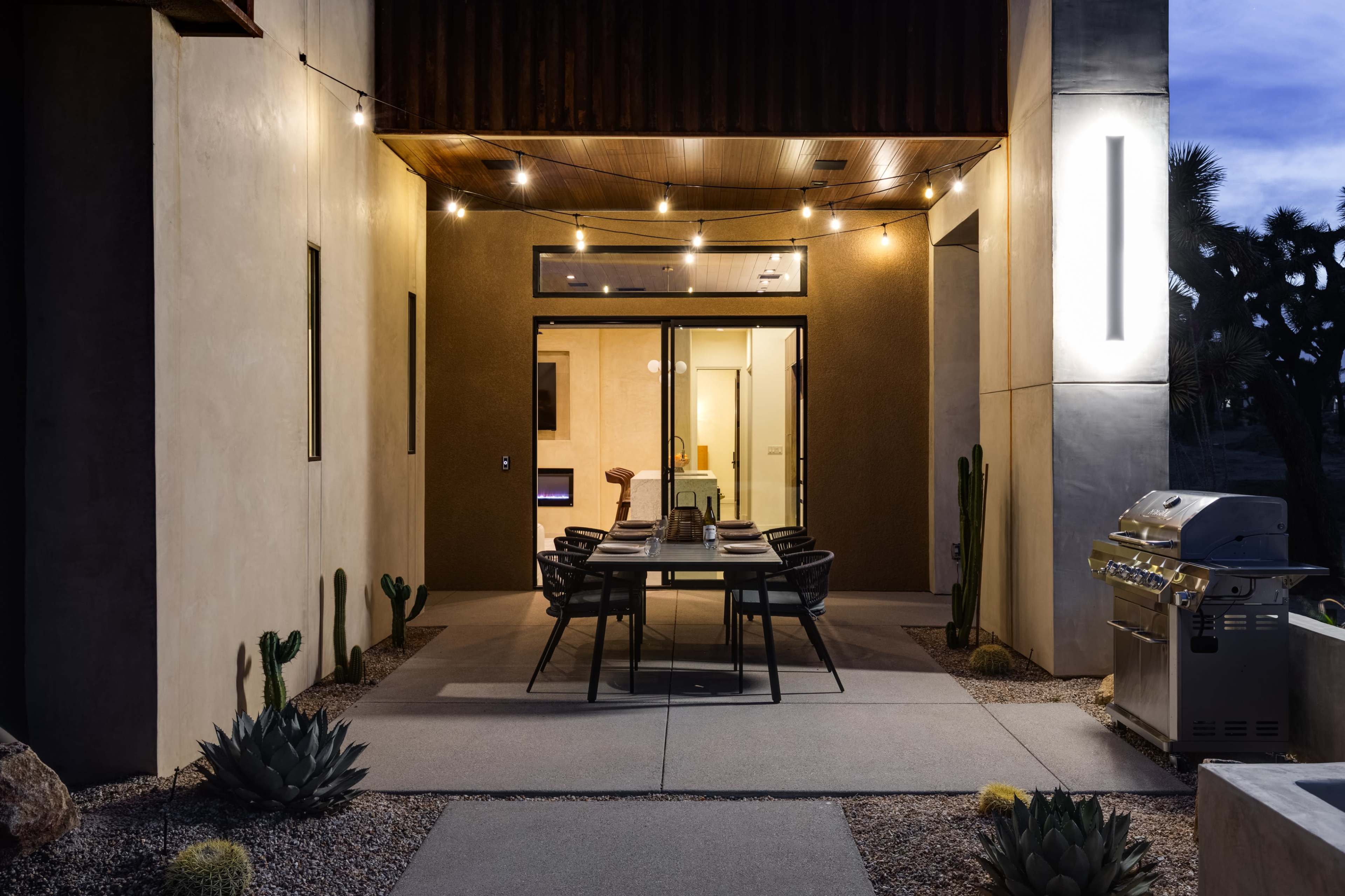 The image shows a modern outdoor dining area with a large table and chairs, string lights overhead, and a grill nearby, surrounded by desert landscaping.