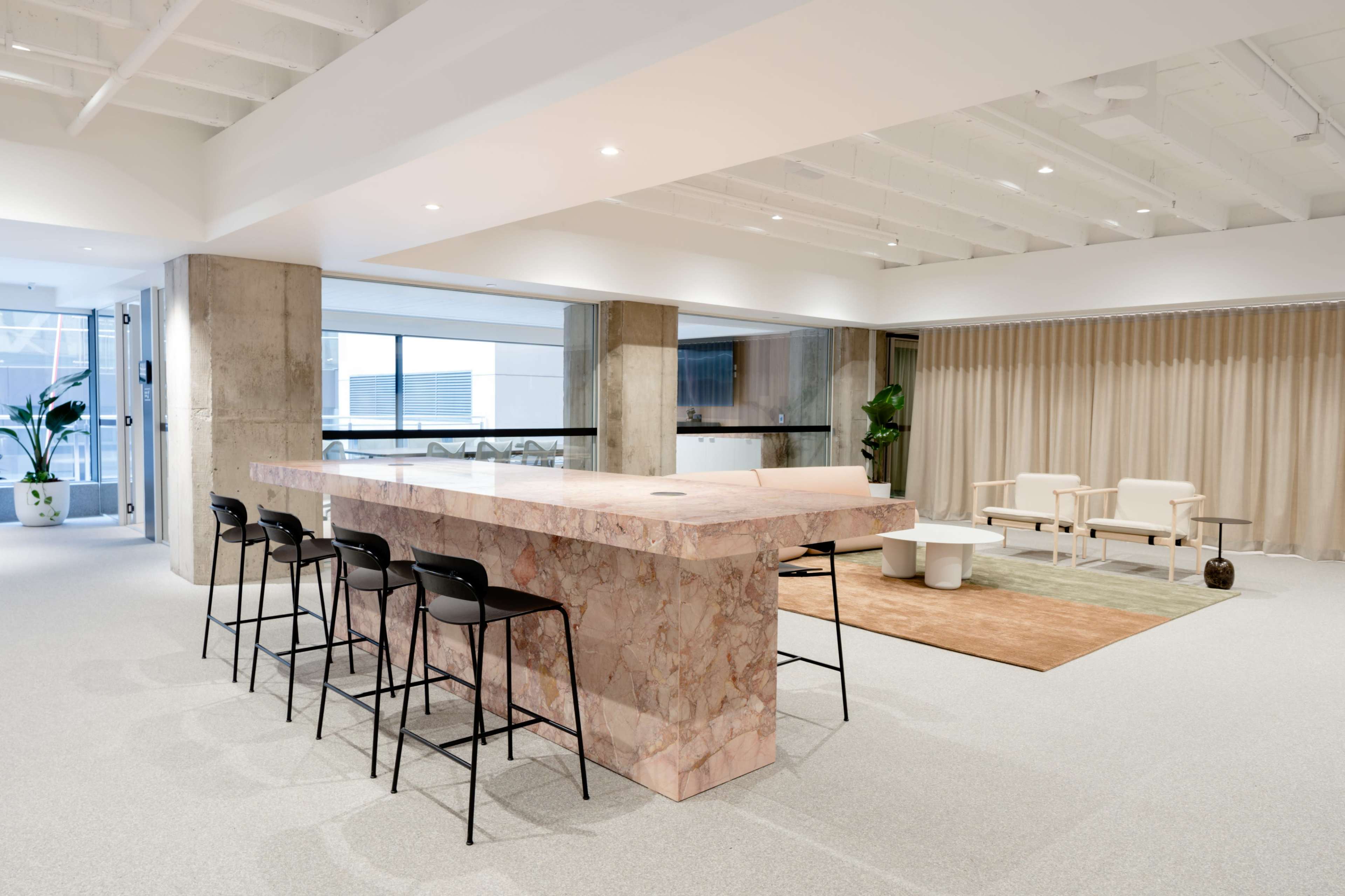 A modern interior space featuring a large pink marble countertop with black bar stools, a seating area with beige chairs, and a light-colored rug on the floor.