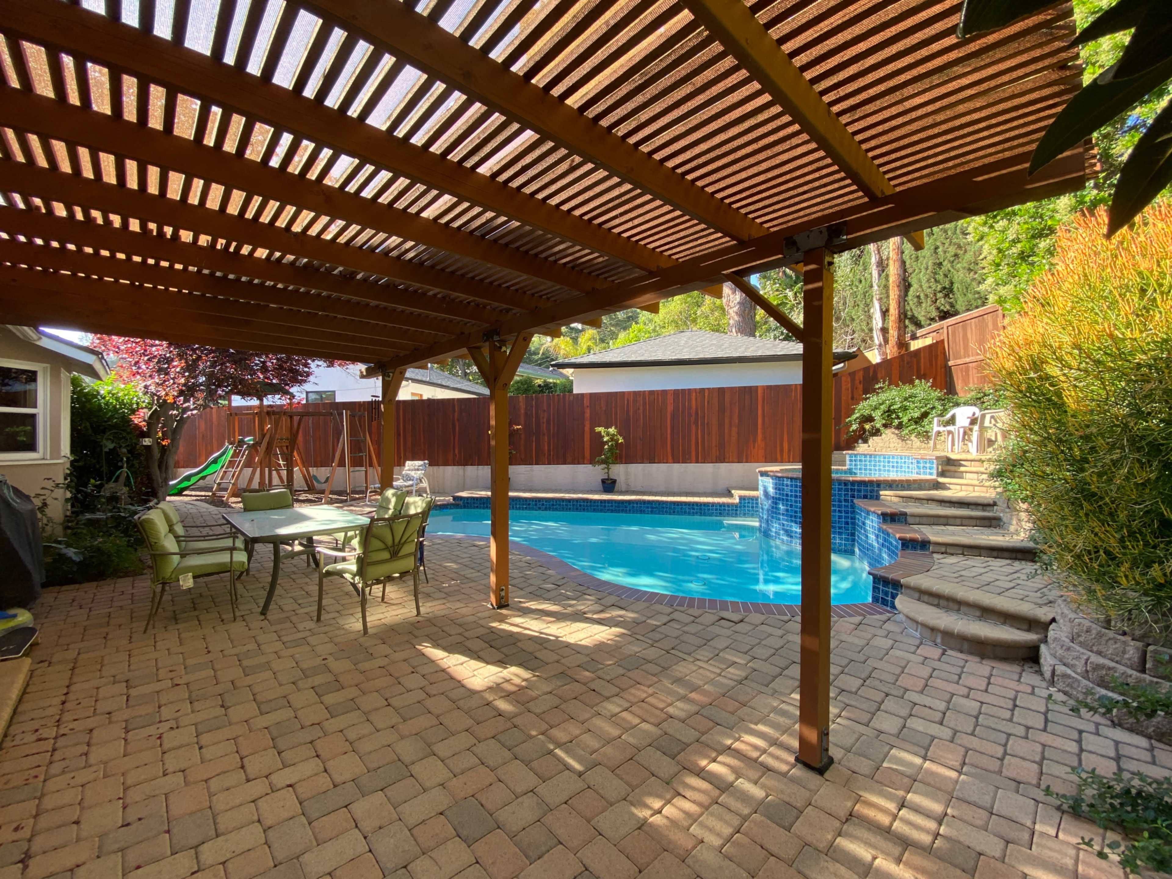 A backyard features a wooden pergola over a patio with a table and chairs, next to a swimming pool surrounded by landscaping.