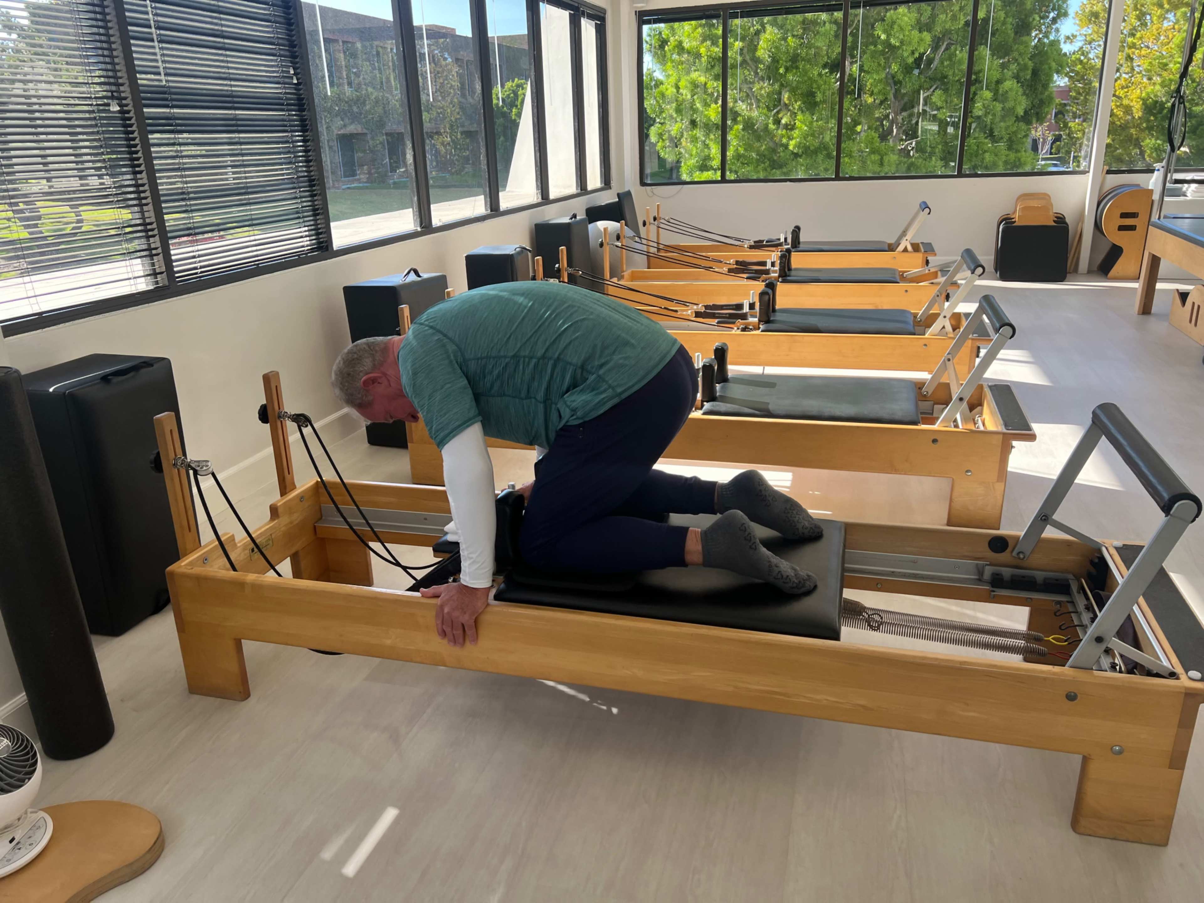 A man is practicing Pilates on a reformer machine in a well-lit exercise studio with several other reformers in the background.