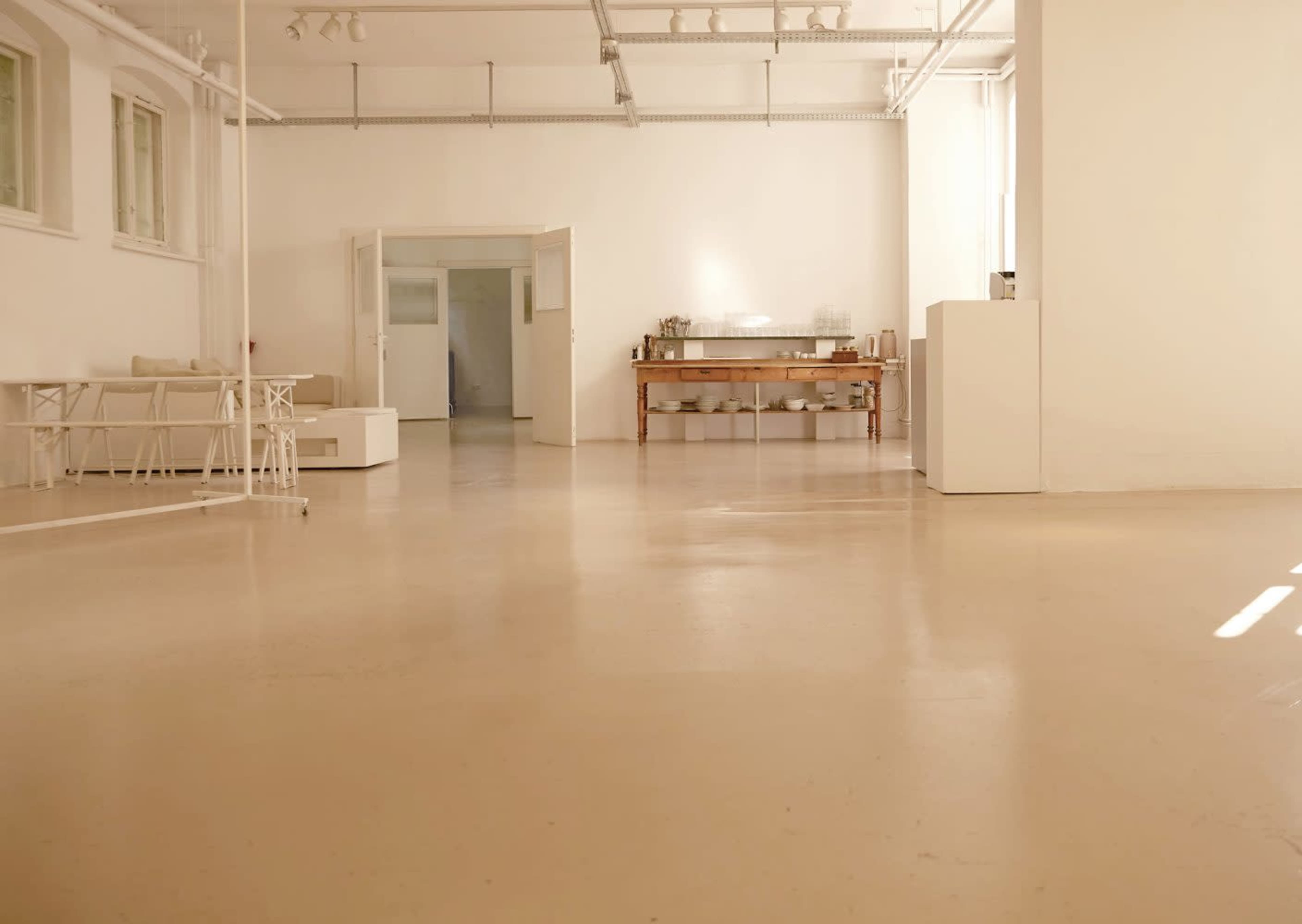 The image shows a spacious room with light-colored walls and floors, featuring a seating area and a table set against one wall.