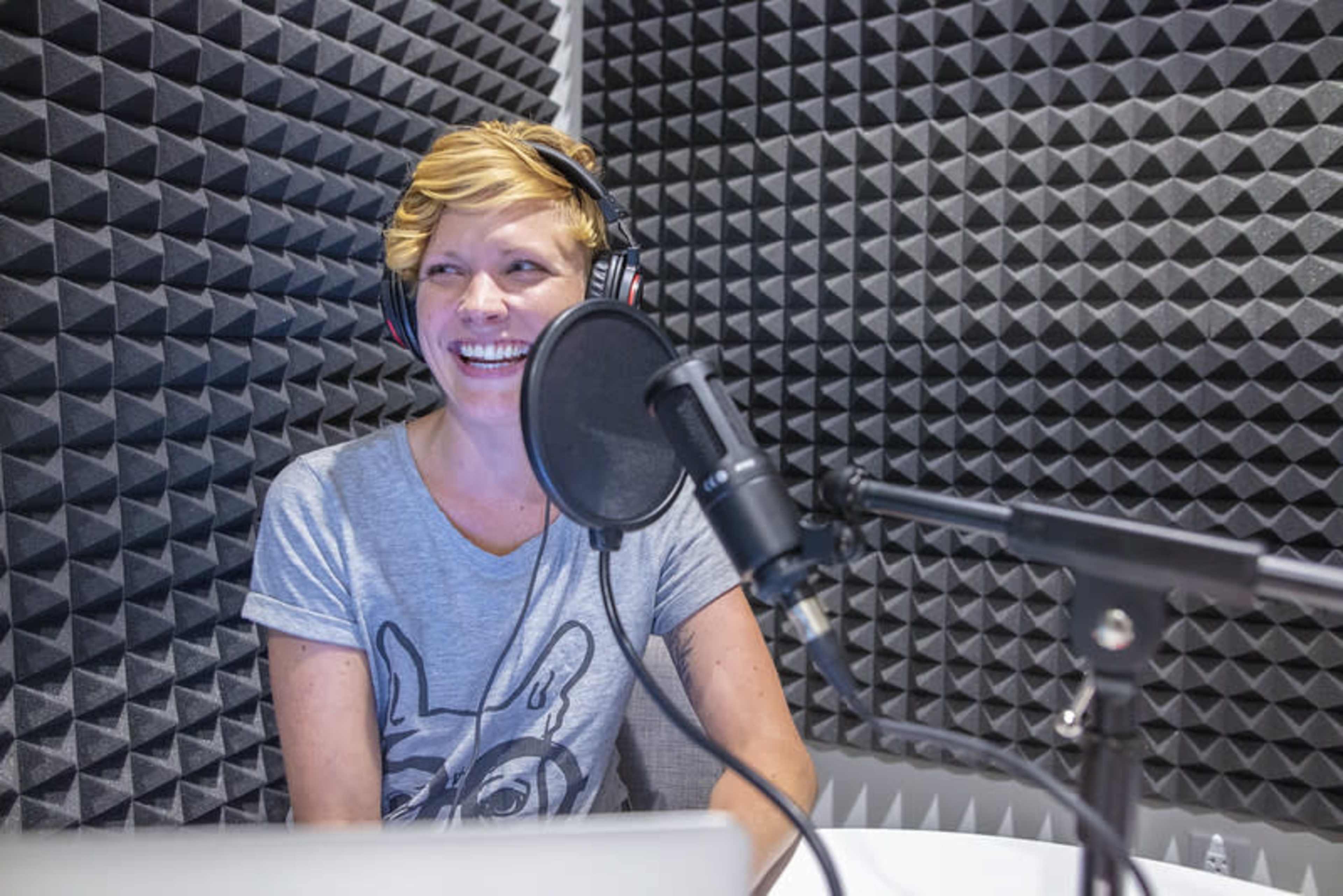 A person with short blonde hair wearing a T-shirt sits in a recording booth equipped with soundproofing panels and a microphone.