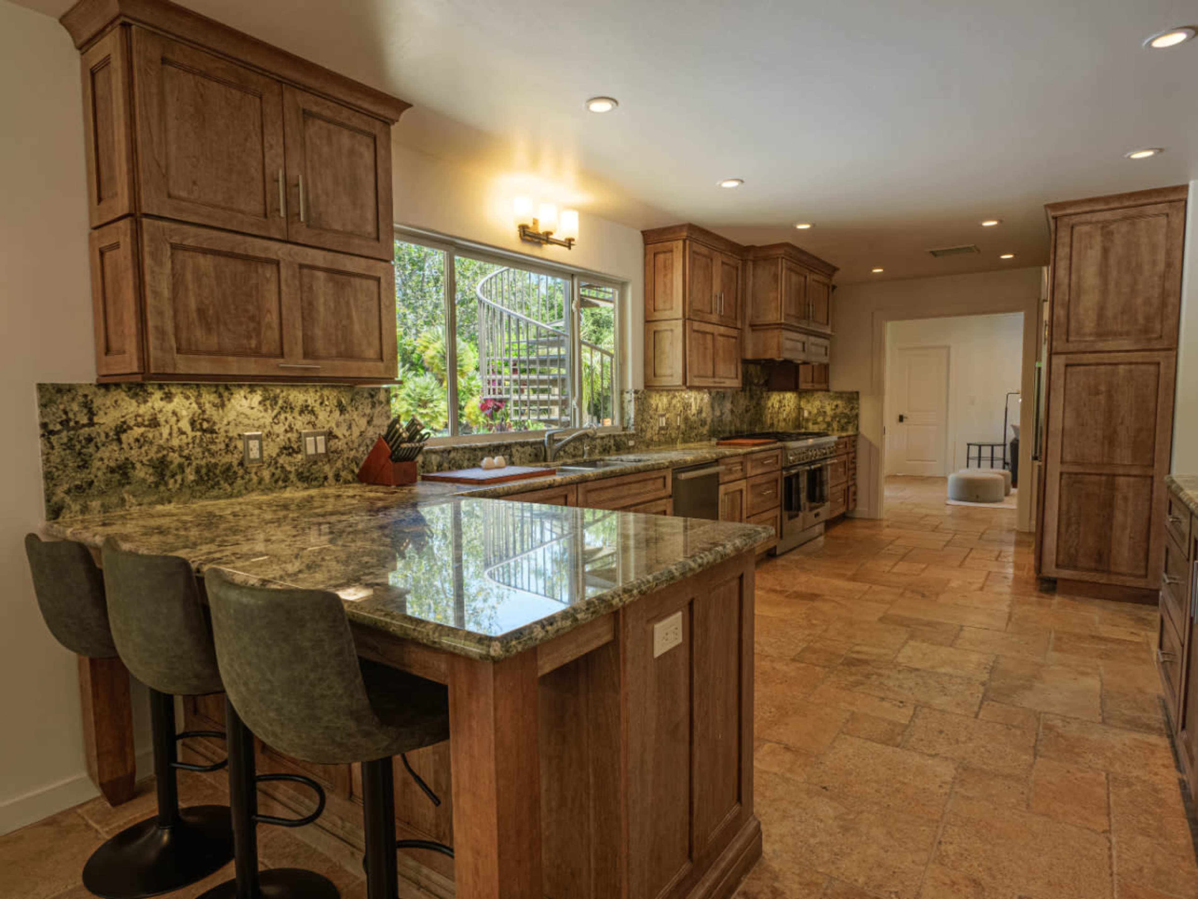 The kitchen features wooden cabinets, granite countertops, and a large window overlooking a garden.