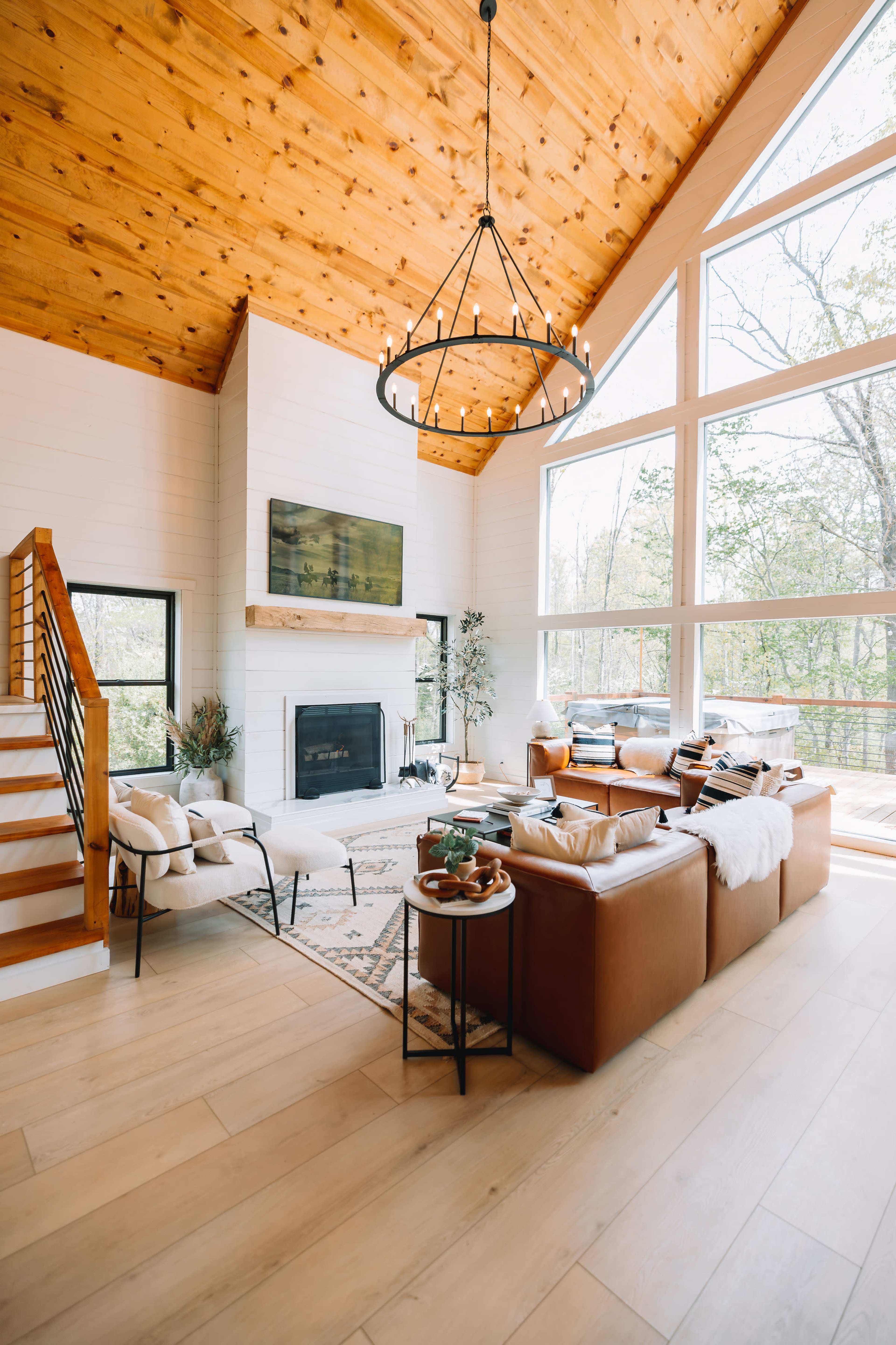 A modern living room features large windows, a fireplace, and a combination of leather sofas and minimalist furniture under a wood-paneled ceiling.