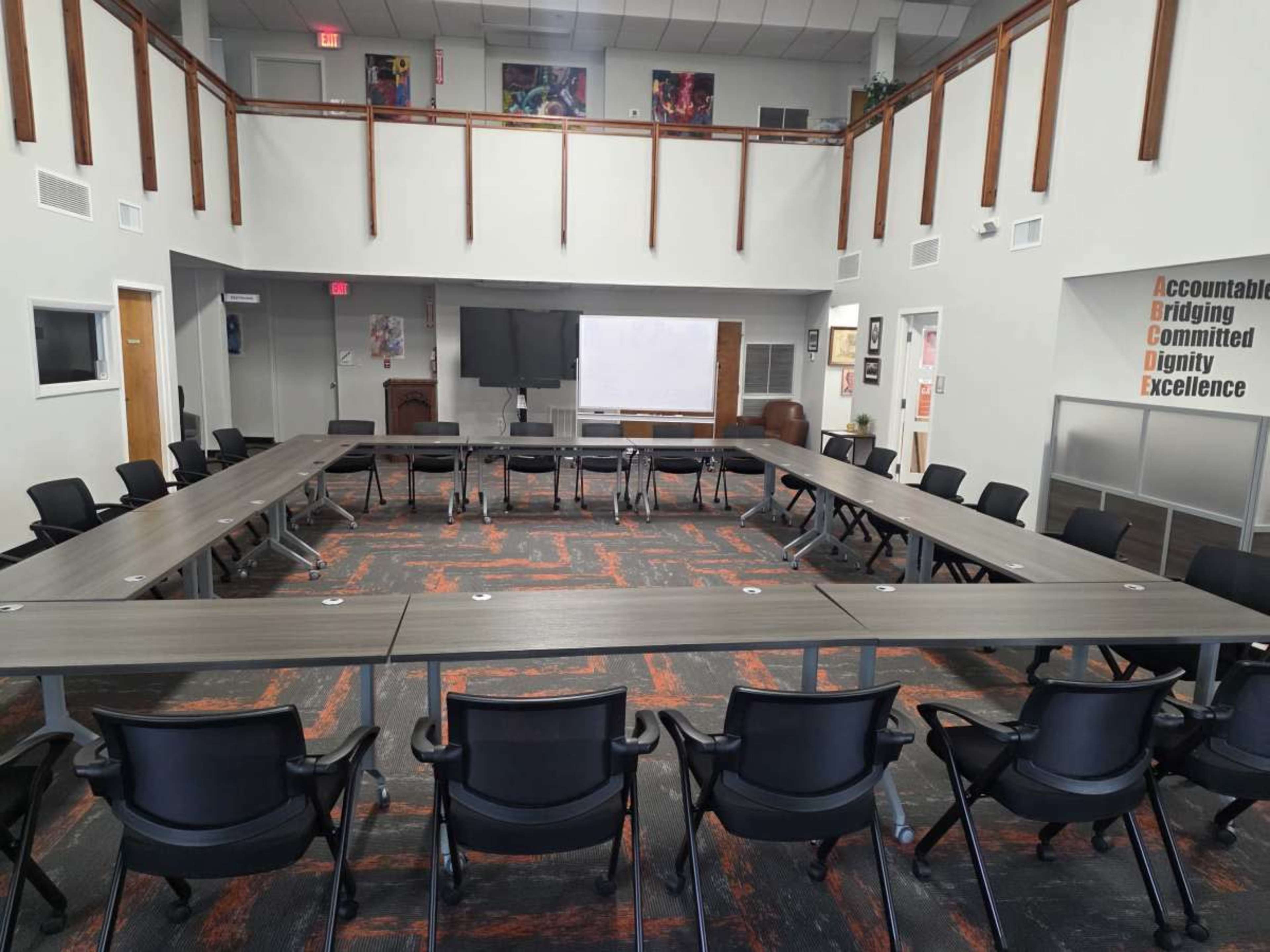 The image shows a conference room arranged with a large rectangular table surrounded by black chairs, flanked by large windows and various wall decorations.
