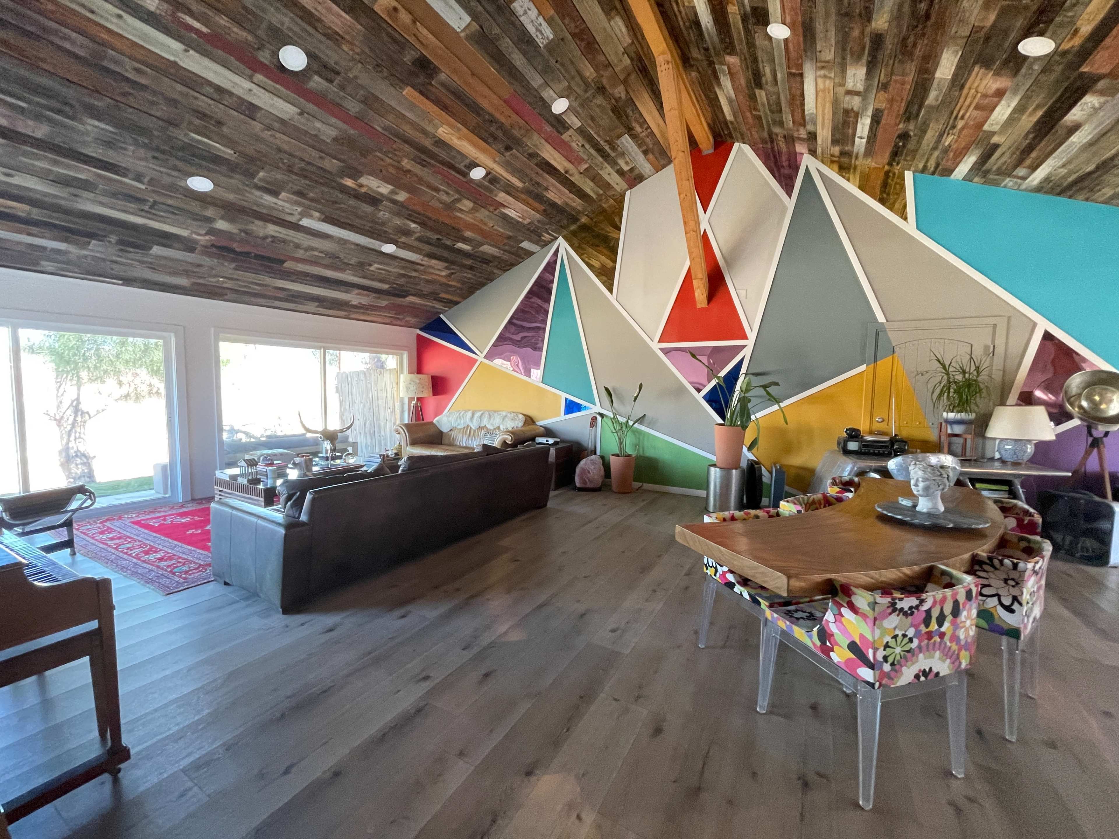 The interior features a modern living space with a painted geometric accent wall, a mix of colorful furniture, and large windows allowing natural light.