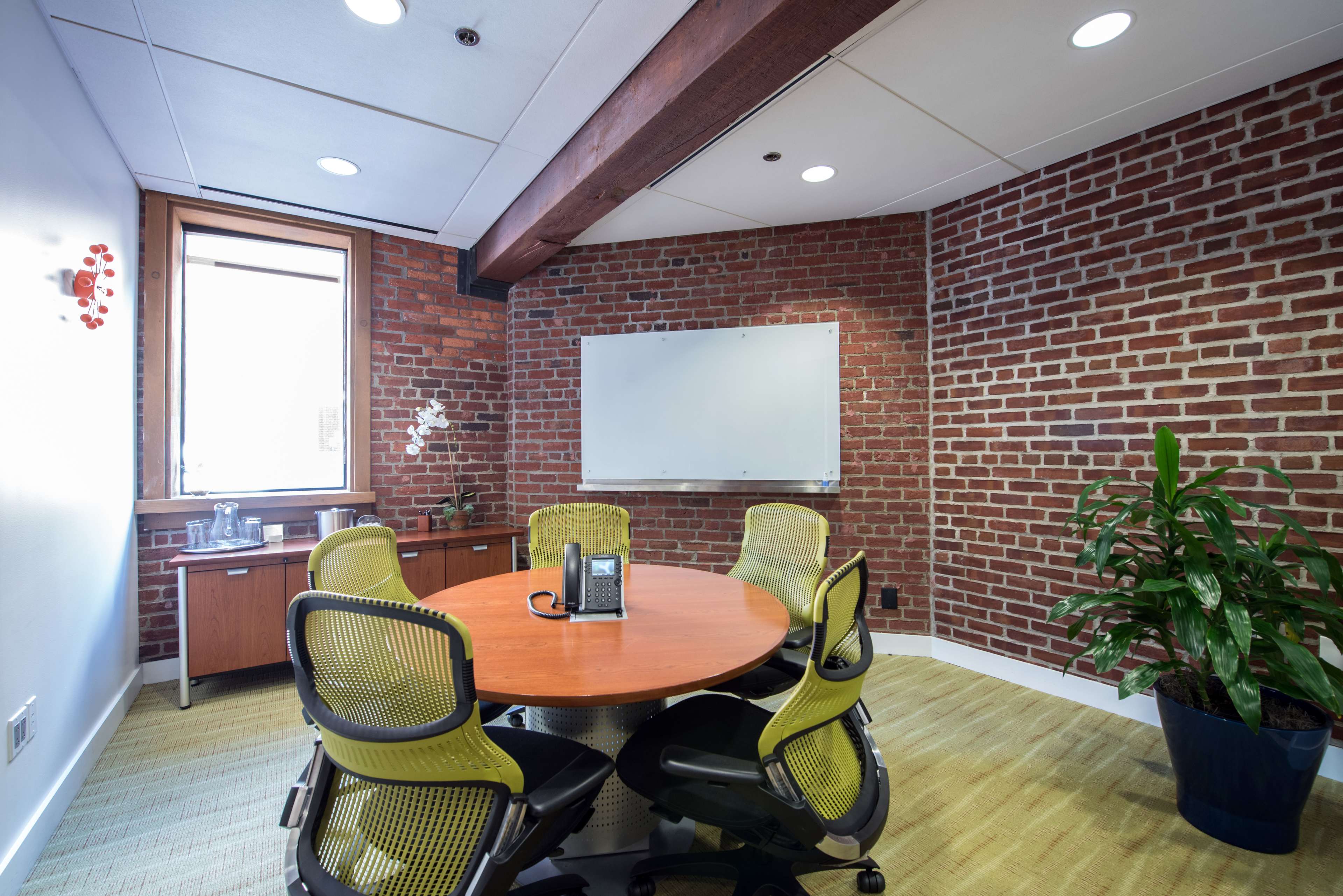 A round wooden table surrounded by green office chairs is positioned in a meeting room with exposed brick walls and a whiteboard.