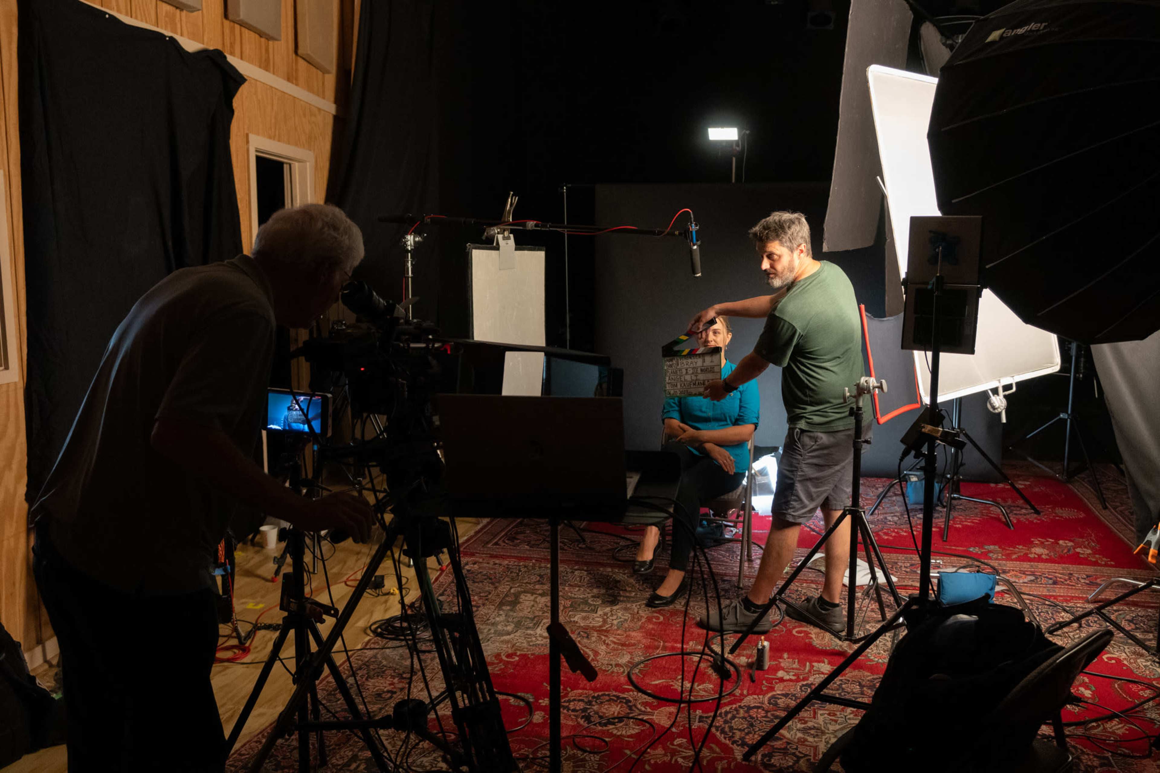 A film crew sets up a shot in a studio with a woman sitting on a chair under bright lights and a camera focused on her.
