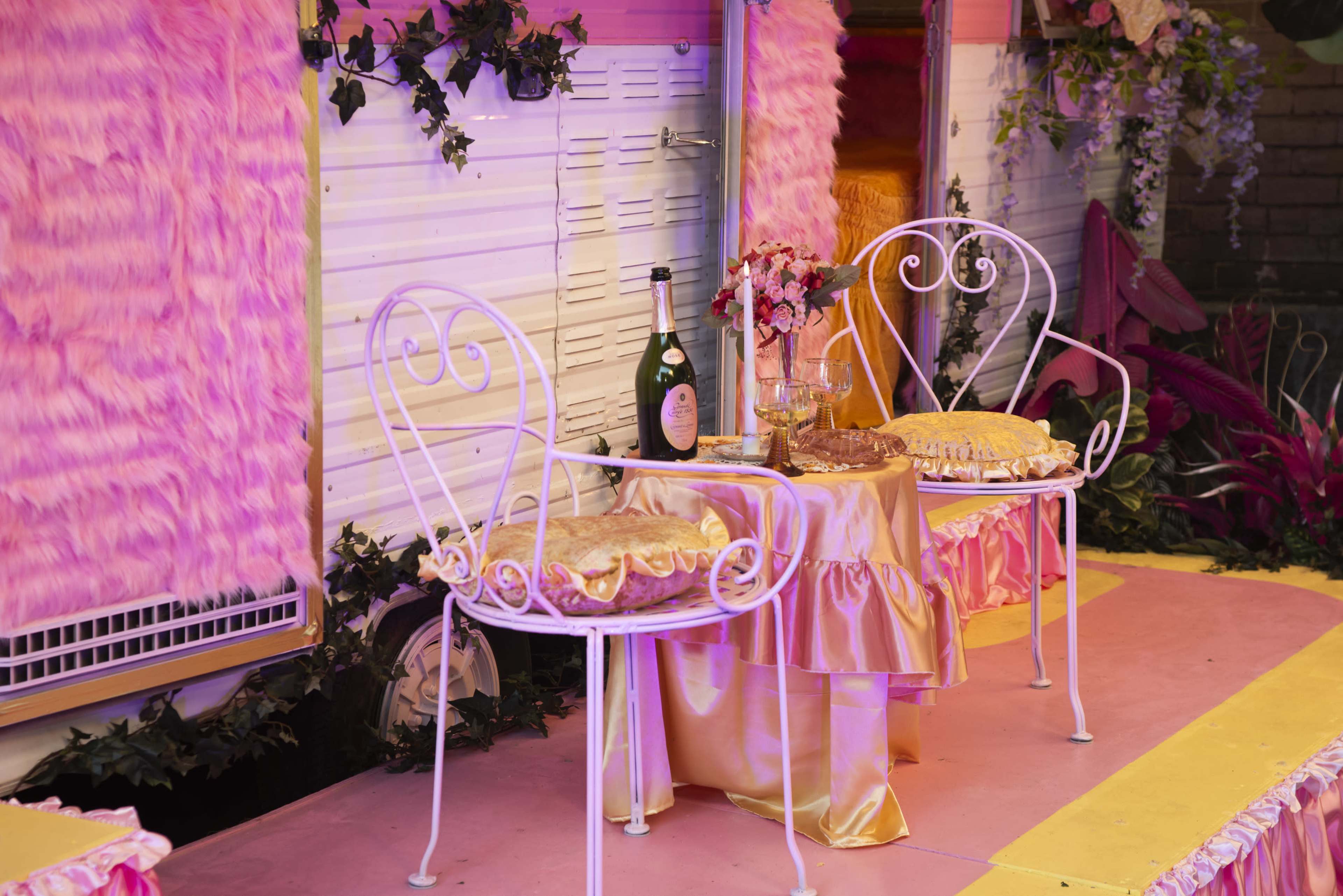 A decorated outdoor space features two white metal chairs with padded cushions beside a small table adorned with a pink tablecloth, a decorative vase, and a bottle of champagne.