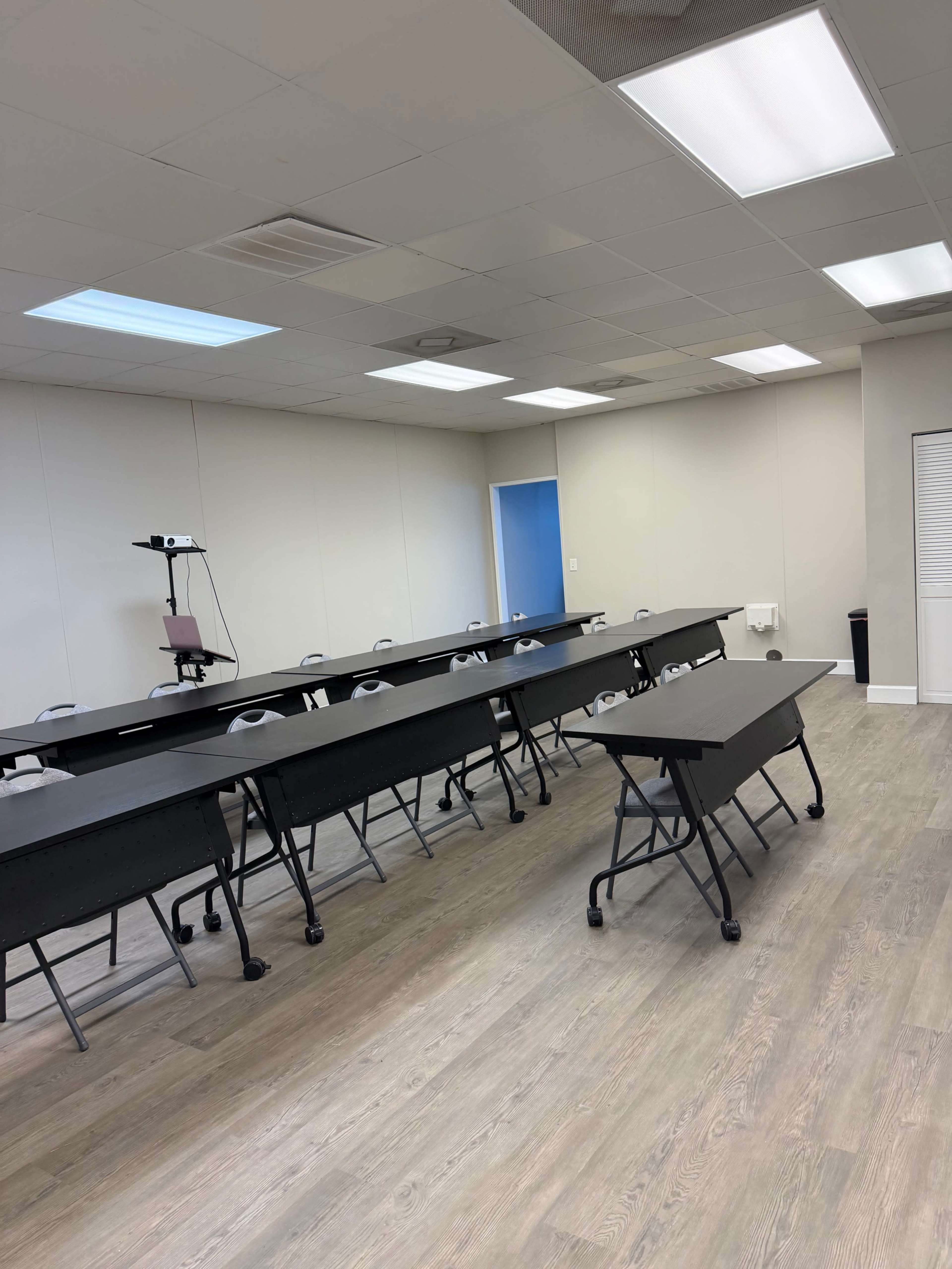 The image shows an empty classroom with multiple black folding tables arranged in rows, illuminated by overhead fluorescent lights.