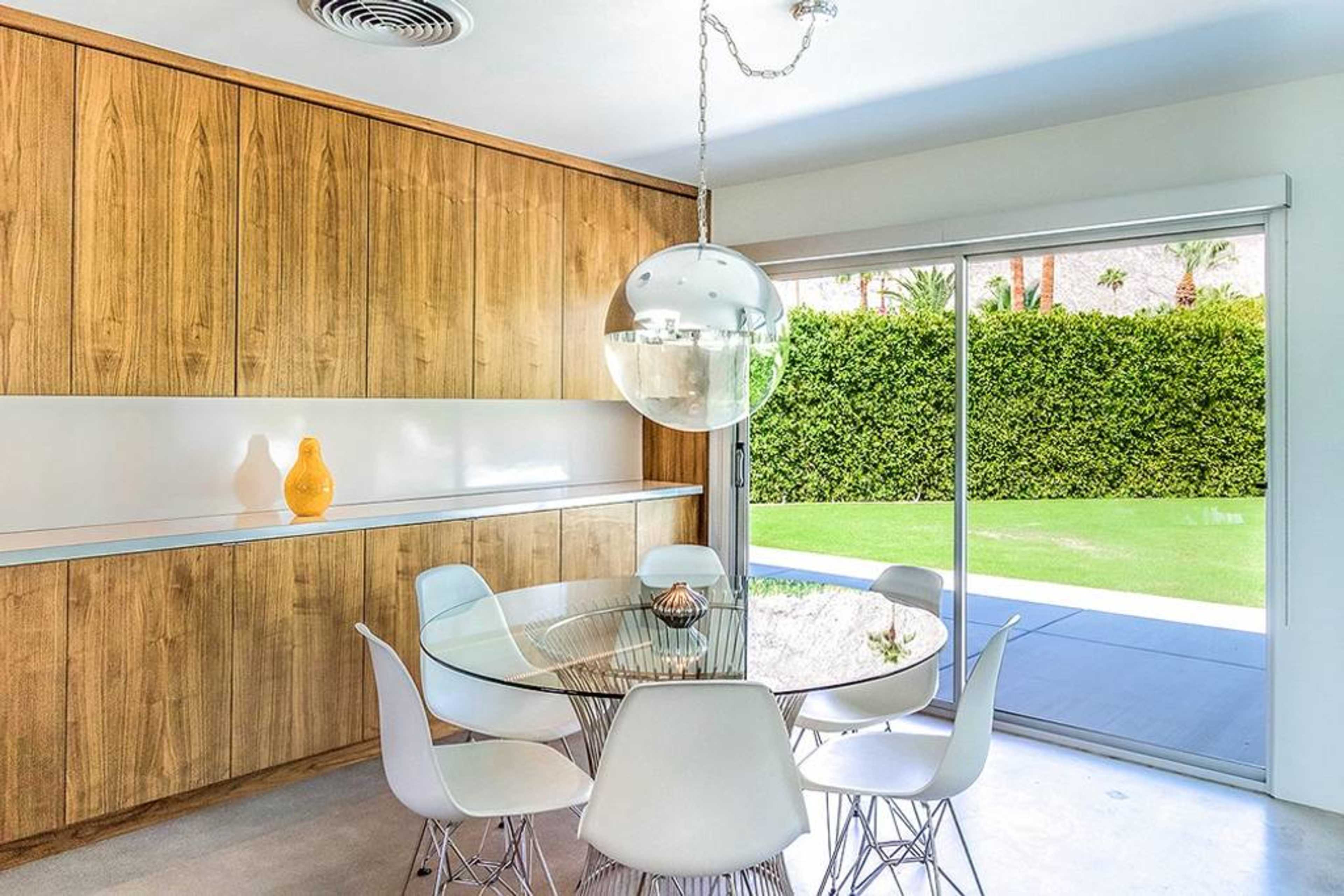 A modern dining area features a round glass table surrounded by white chairs, with a wooden wall unit and large sliding doors leading to a grassy outdoor space.