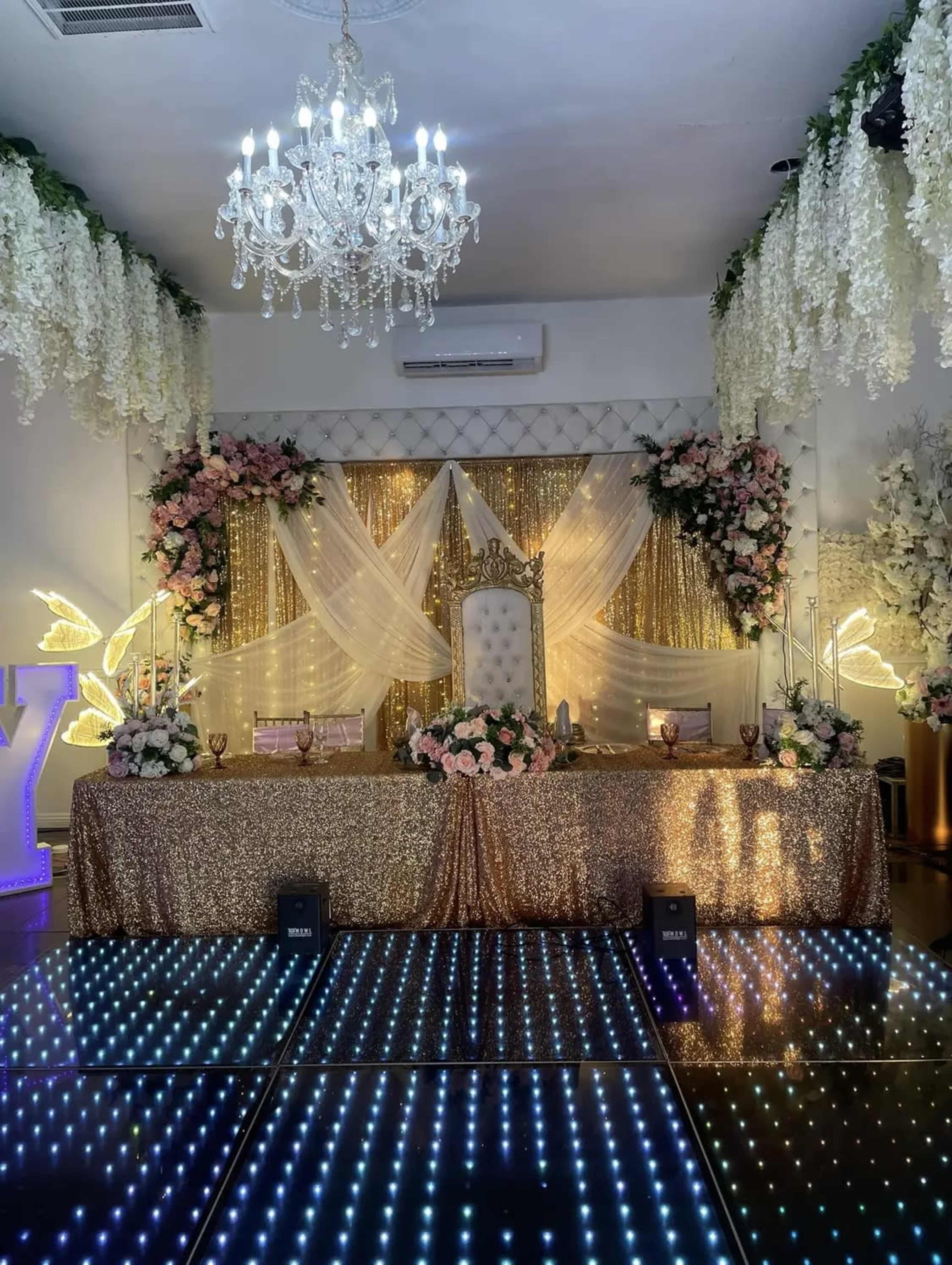 The image shows a decorated event space featuring a glittering gold table, a backdrop of draped fabric and flowers, and a chandelier overhead.
