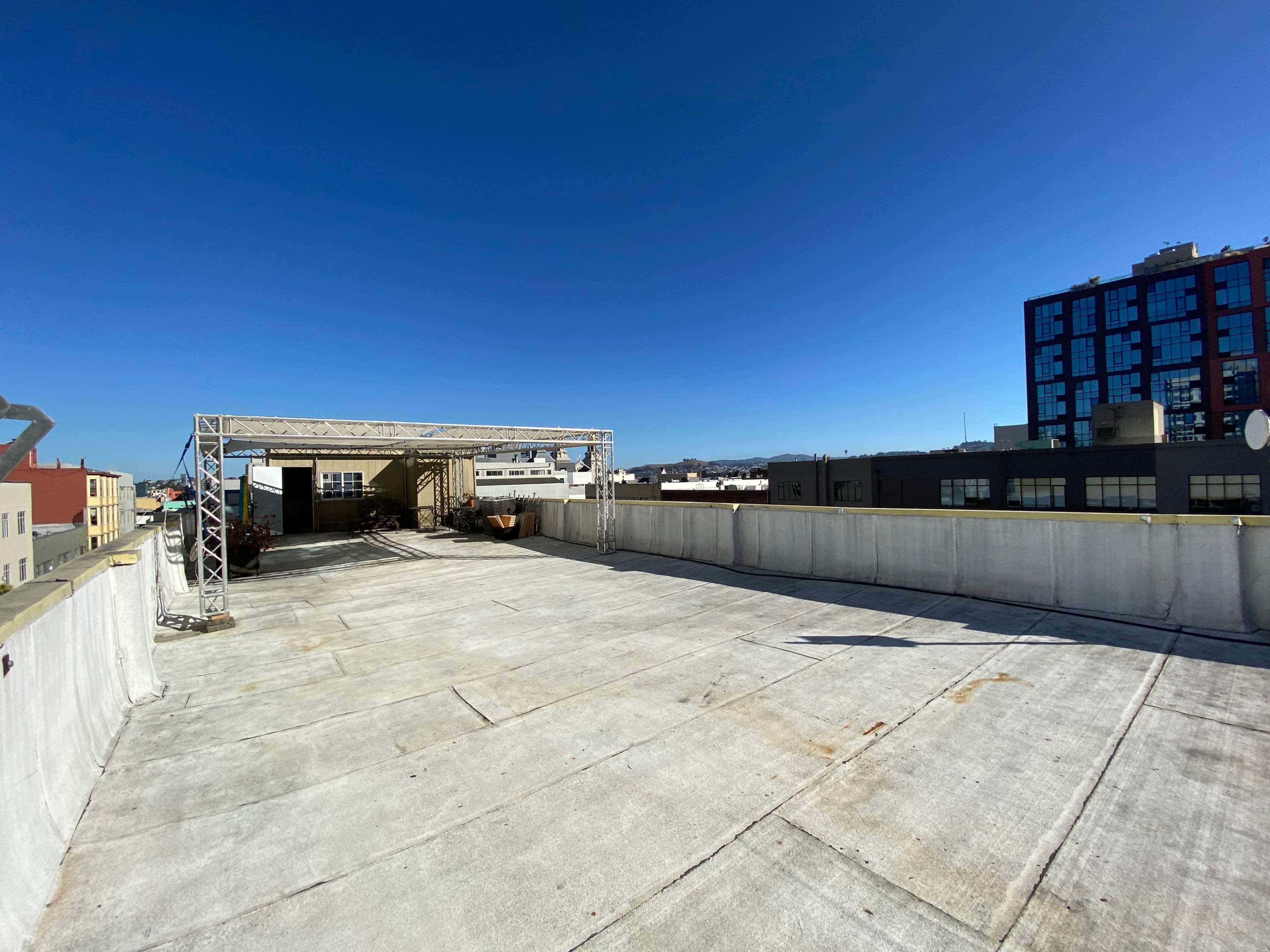 A flat rooftop space with a canopy structure on one side and city buildings in the background under a clear blue sky.