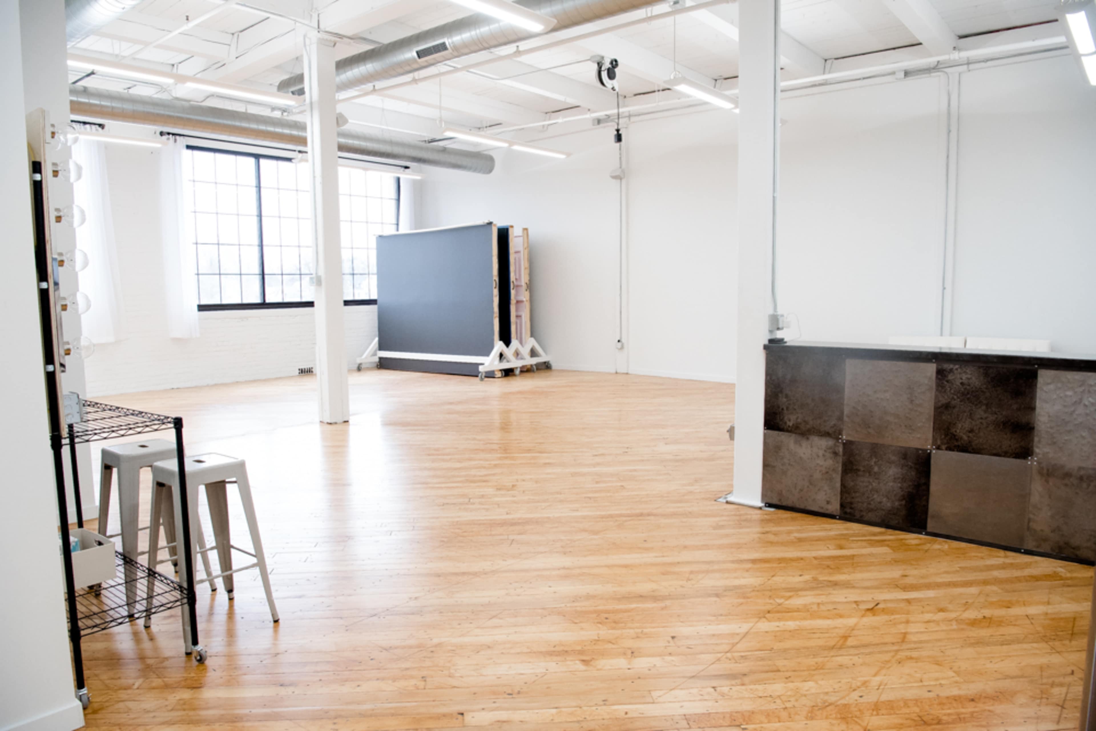 A spacious, empty room with polished wooden floors, large windows, and a minimalist setup including a counter and stools.