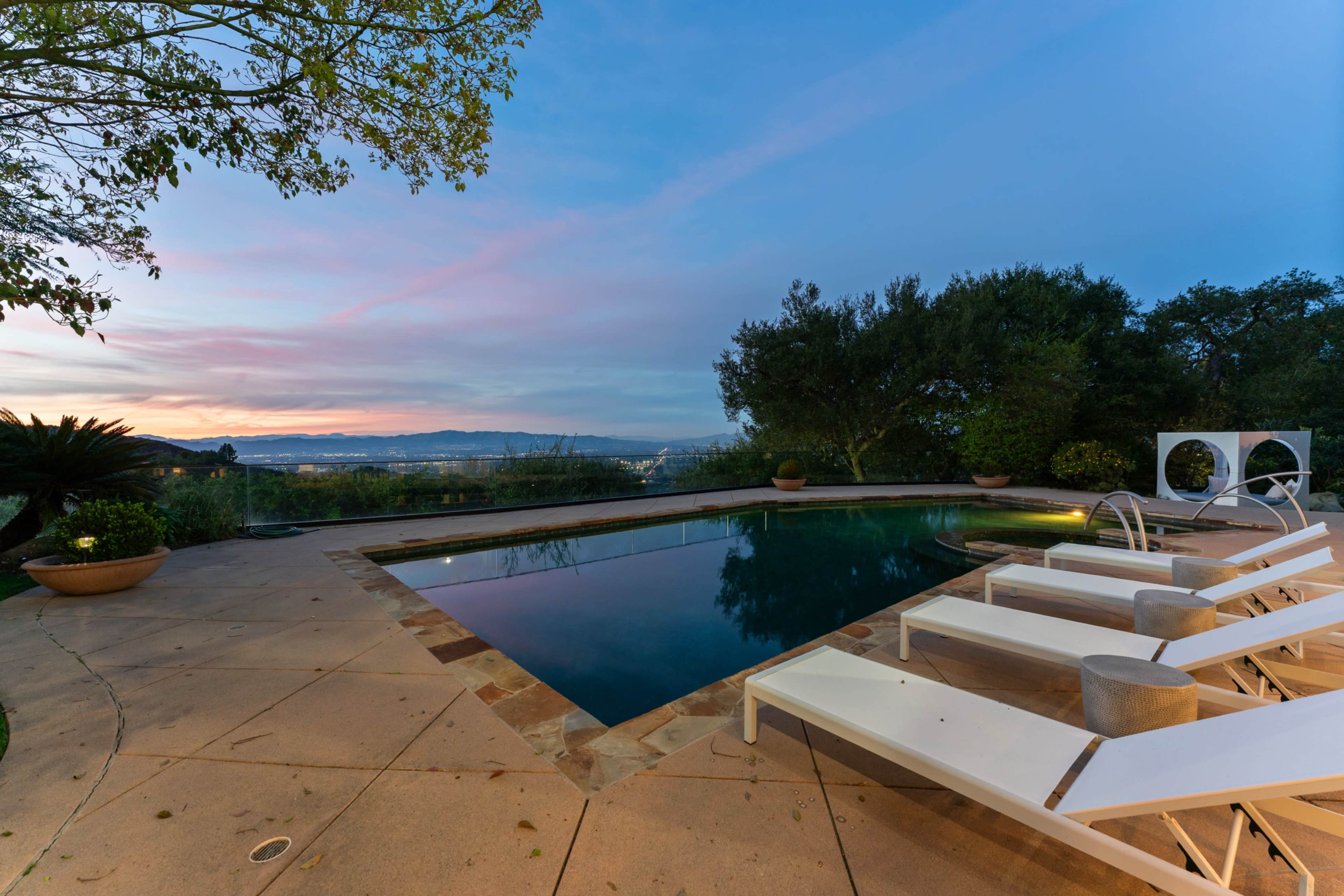 The image shows a swimming pool surrounded by lounge chairs, with a view of lush greenery and a sunset in the background.