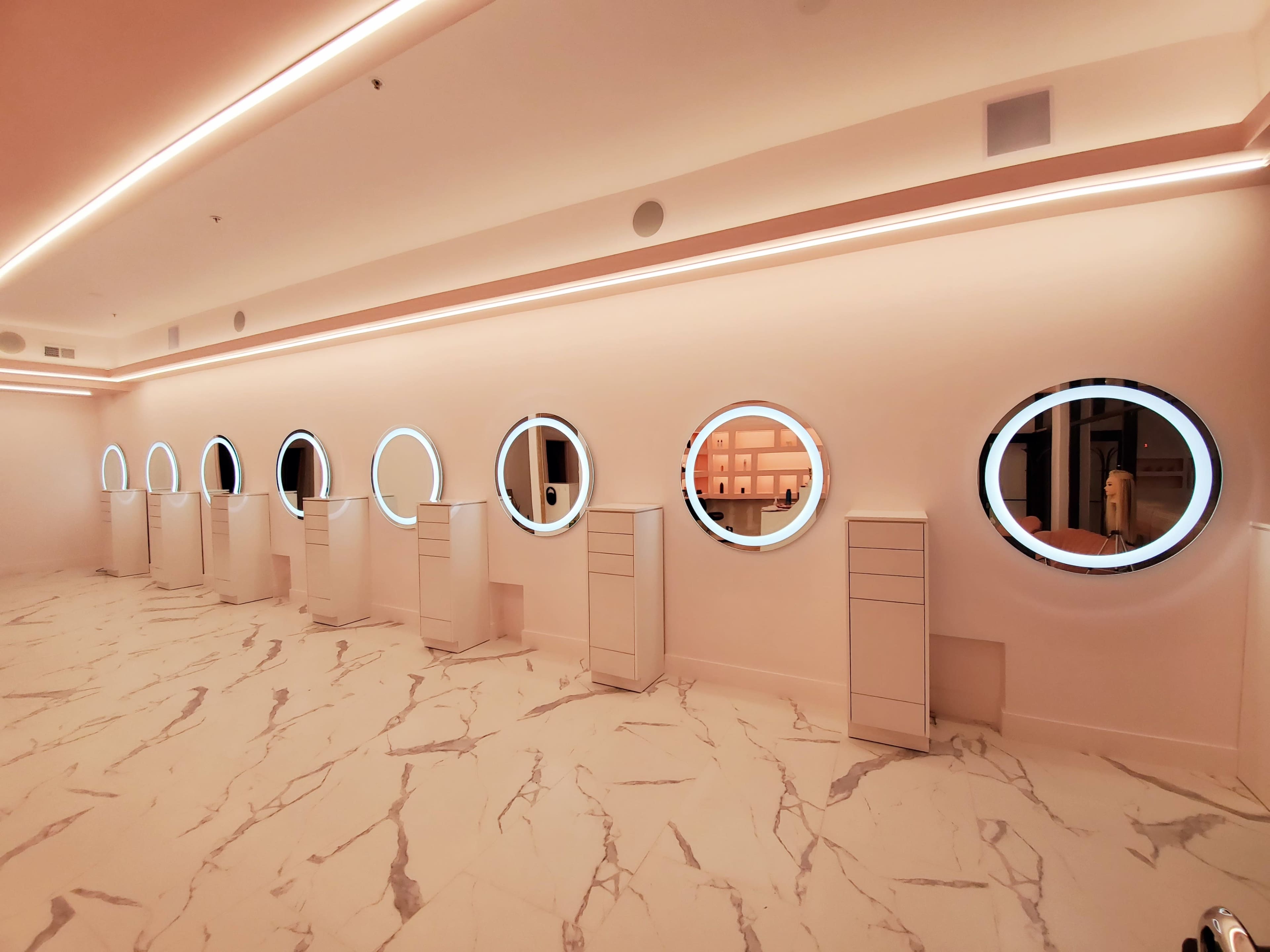 A row of circular mirrors with backlighting mounted on a wall, beside white cabinetry, in a well-lit space with a marble floor.