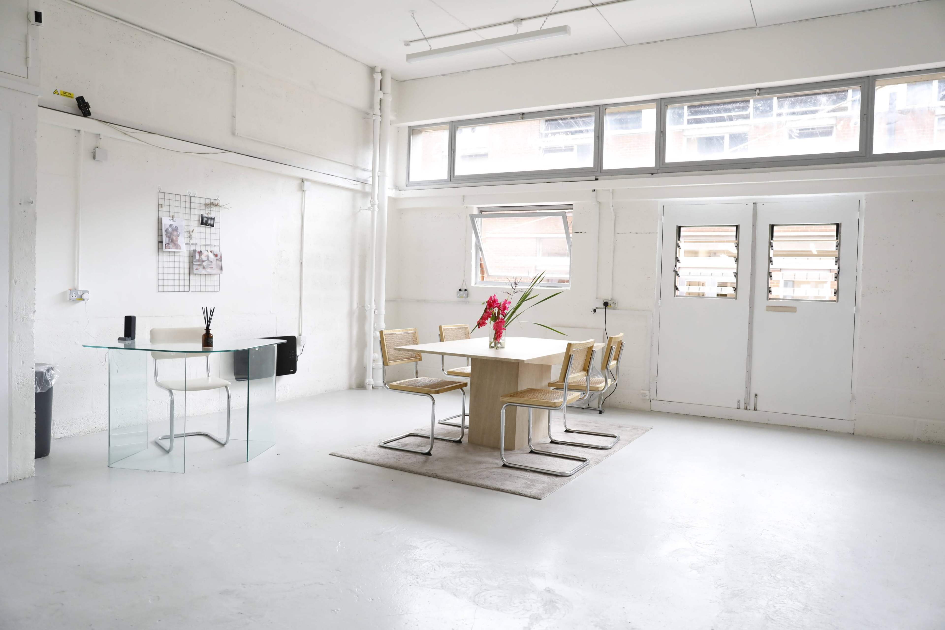A bright, spacious room featuring a glass table and wooden dining table surrounded by chairs, with large windows letting in natural light.