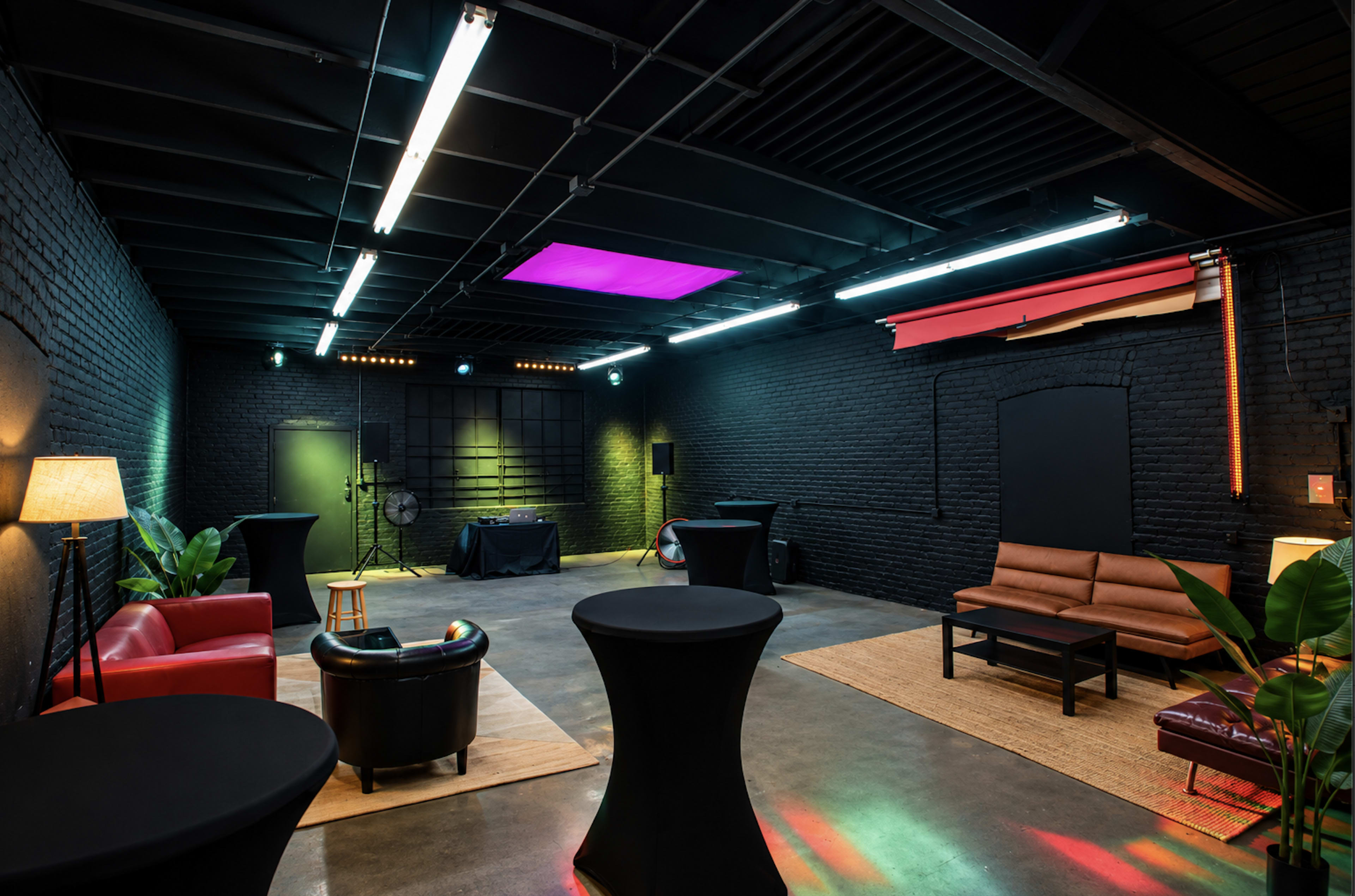 The image shows an industrial-style room with dark brick walls, colorful lighting, and various seating arrangements, including a sofa and lounge chairs.