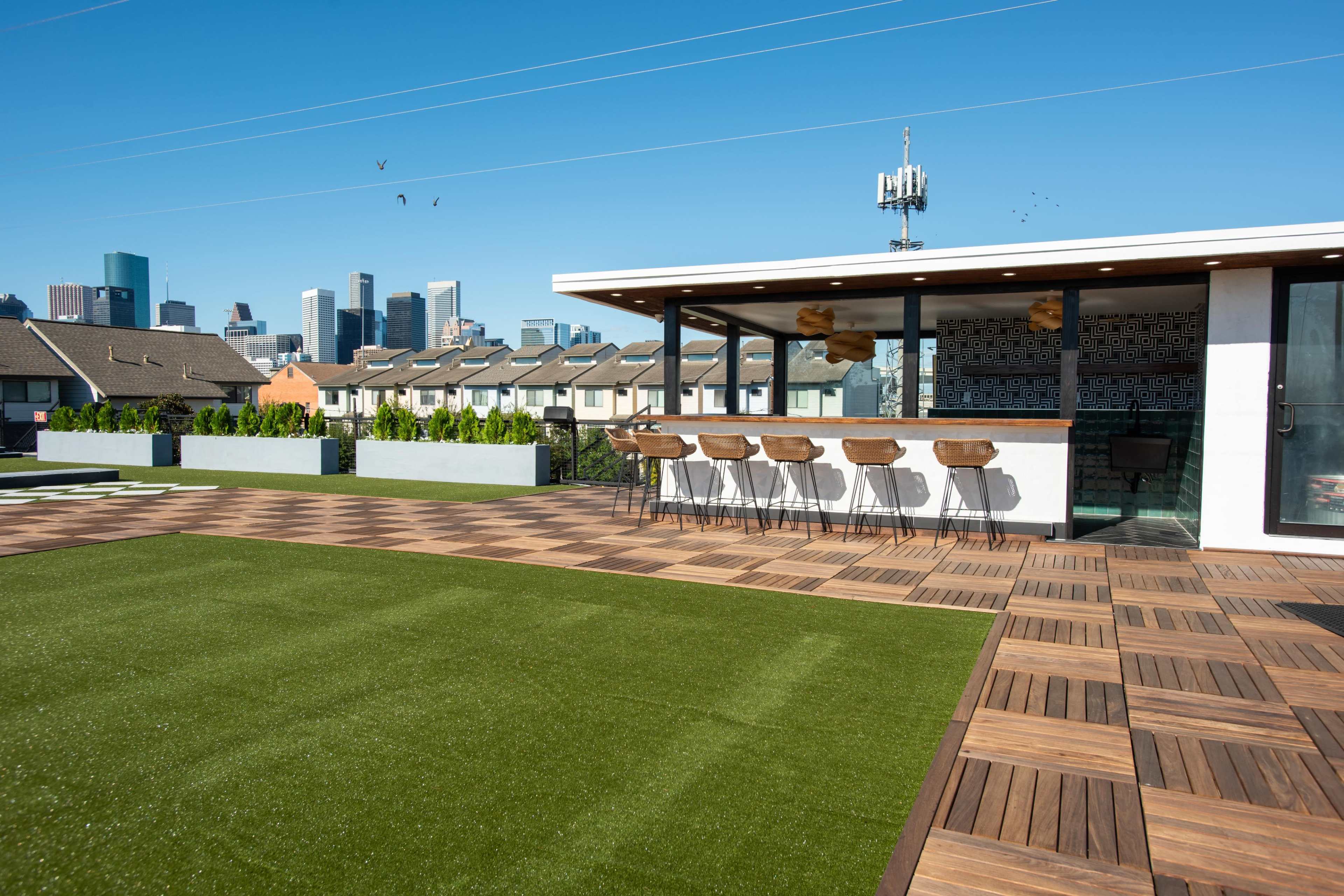 A rooftop terrace featuring a green lawn area, wooden flooring, and a bar counter with high stools, overlooking a city skyline.
