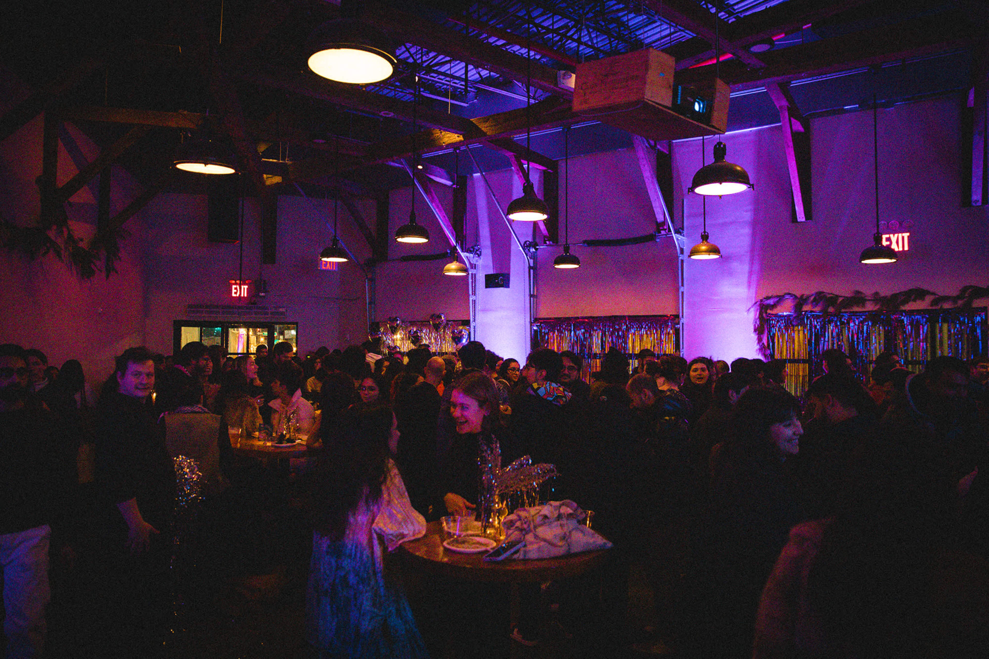 A crowded indoor event features people socializing at tables under purple lighting and industrial-style decor.