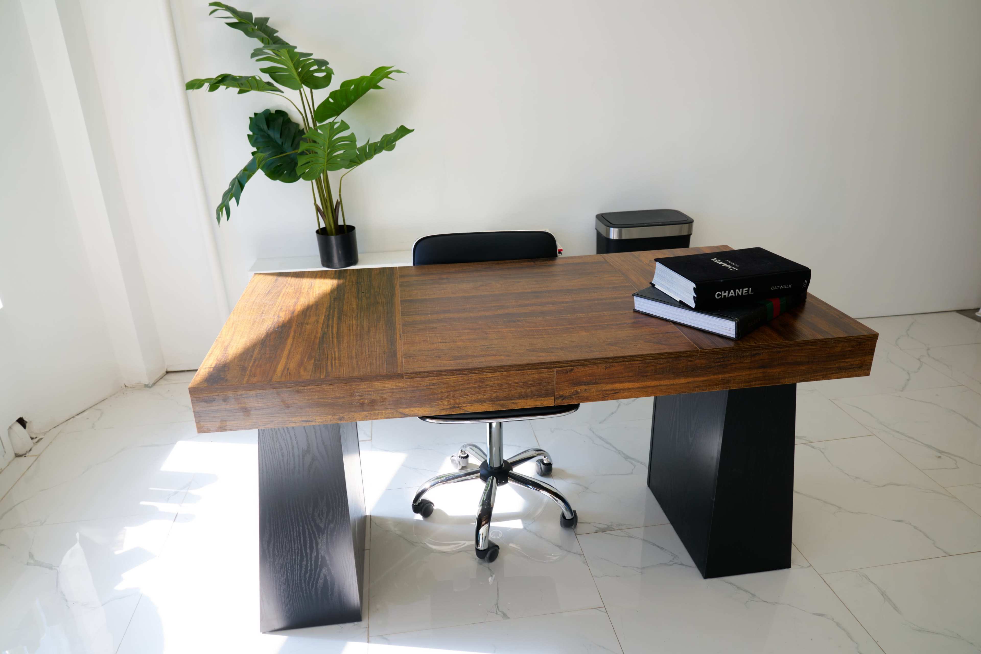 The image shows a contemporary wooden desk with a black office chair, accompanied by a potted plant, and a small trash can in a minimalist room.