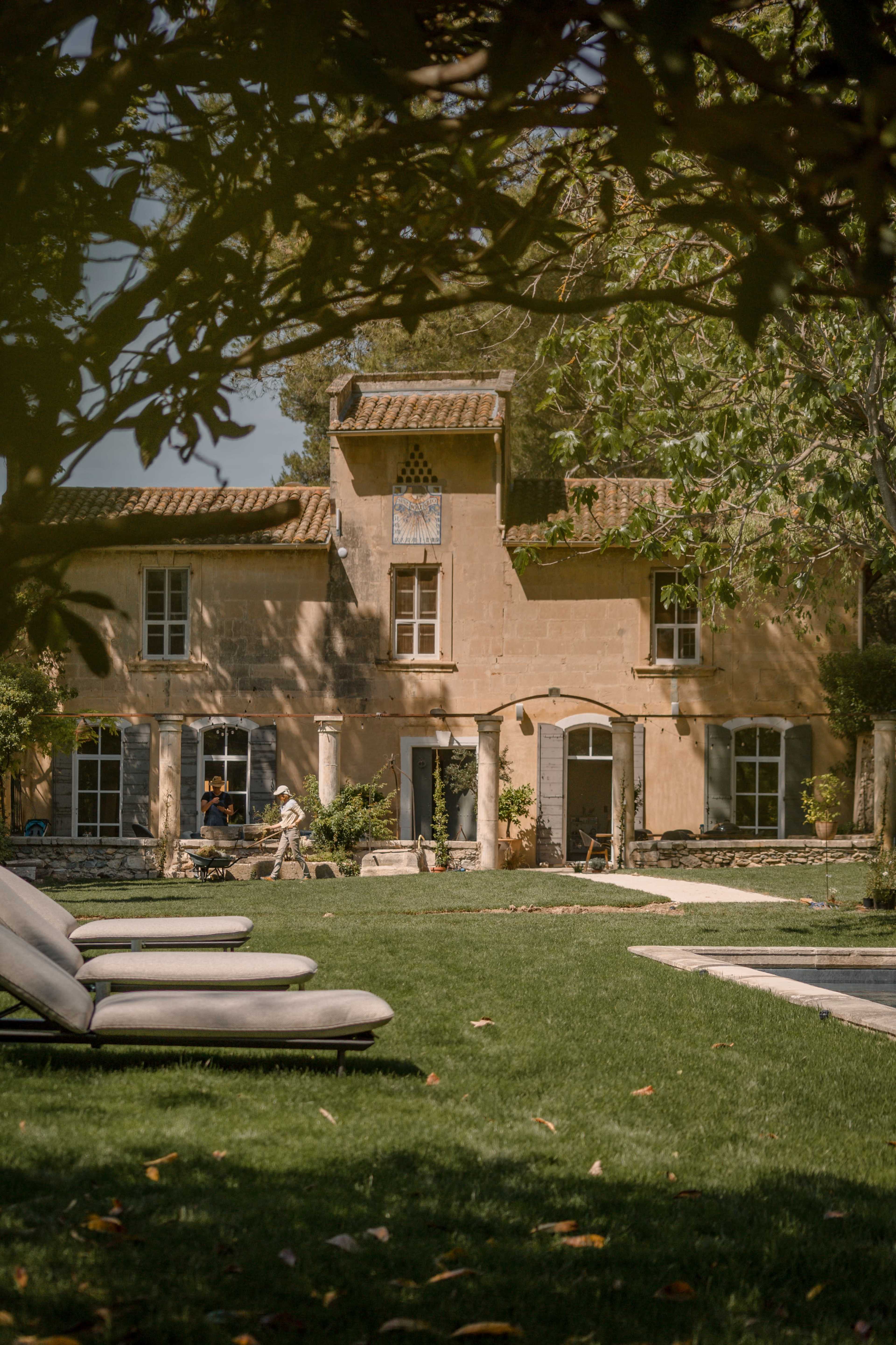 A stone villa with large windows and a tower stands in a lush green yard, surrounded by lounge chairs and trees.