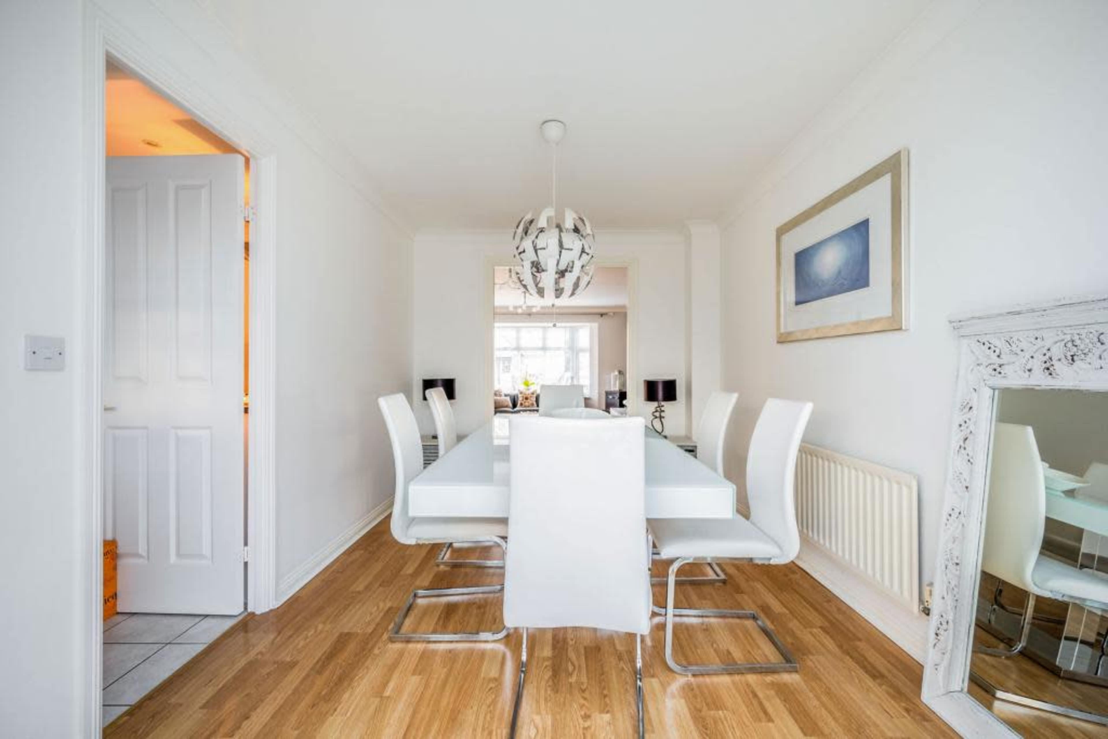 A modern dining room features a white table surrounded by sleek chairs, with light wood flooring and a mirror reflecting the space.