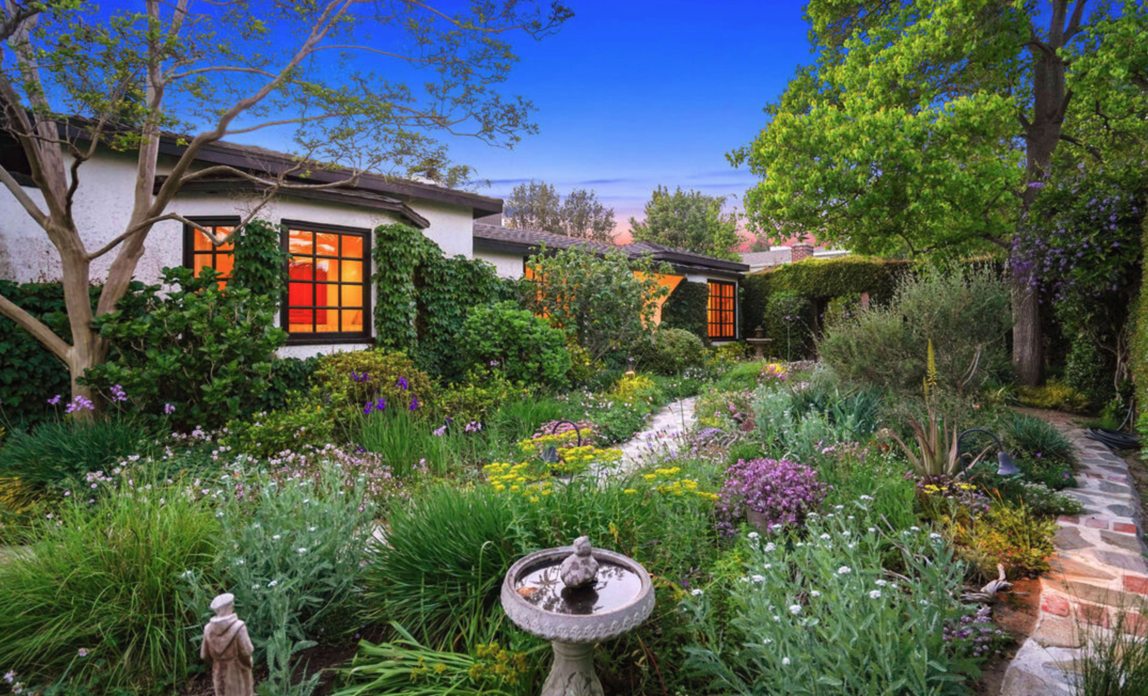 The image shows a lush garden filled with various flowering plants and greenery, featuring a pathway leading to a home with ivy-covered walls and warm orange windows.