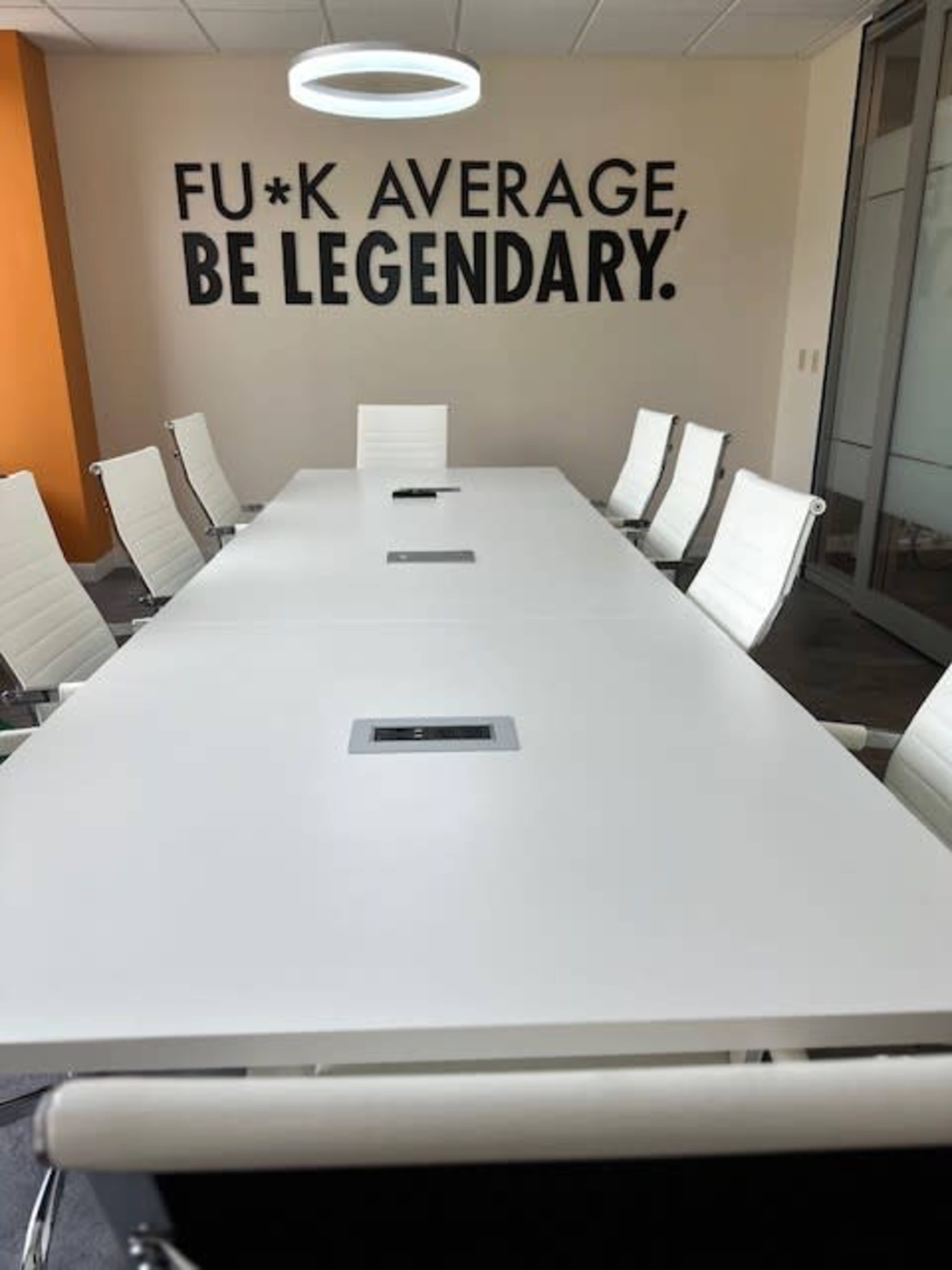 A white conference table with white chairs is set up in a brightly colored meeting room that features a motivational phrase on the wall.