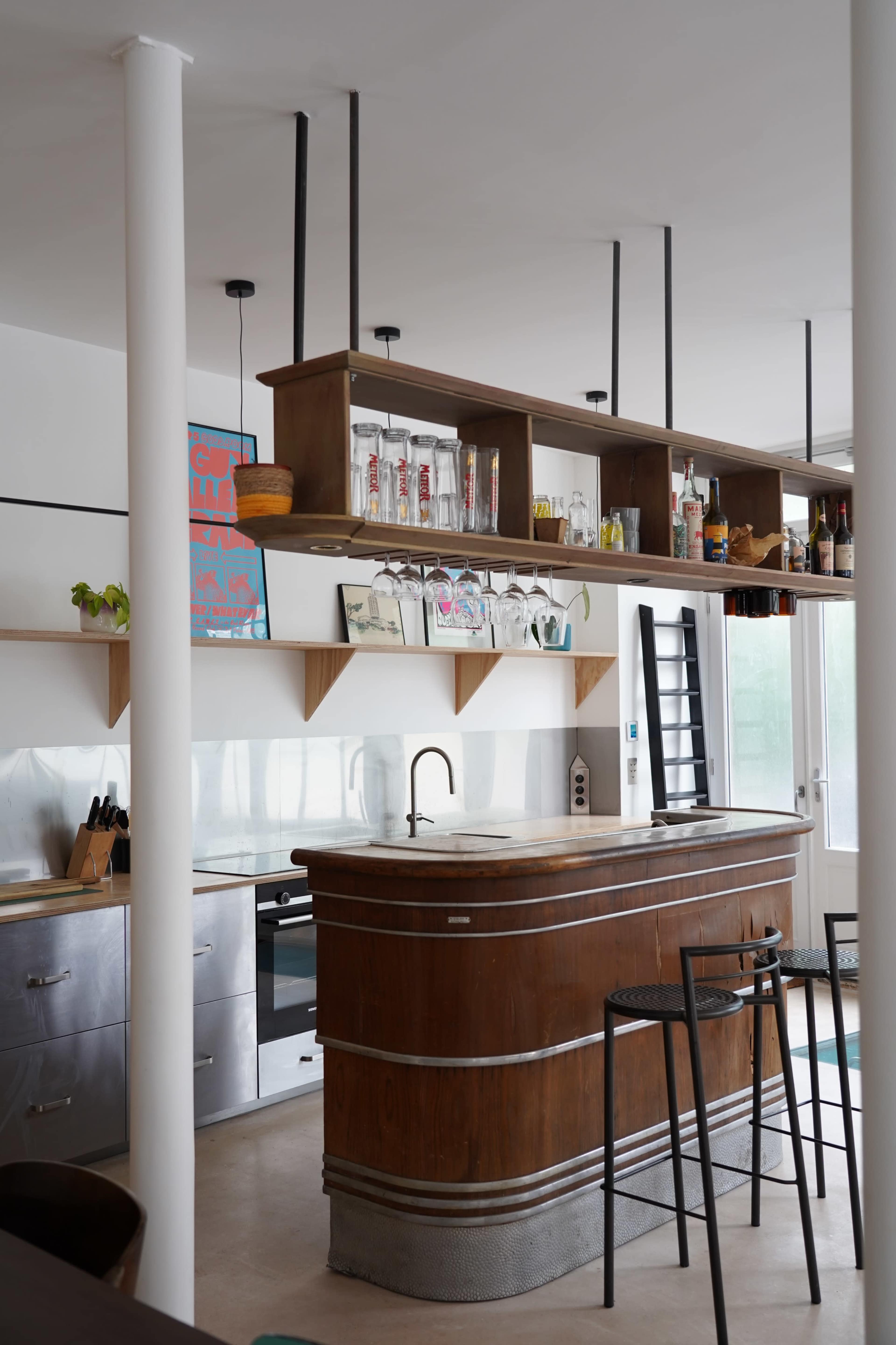 The image shows a minimalist kitchen with a wooden bar counter and overhead shelves displaying various bottles and glassware.
