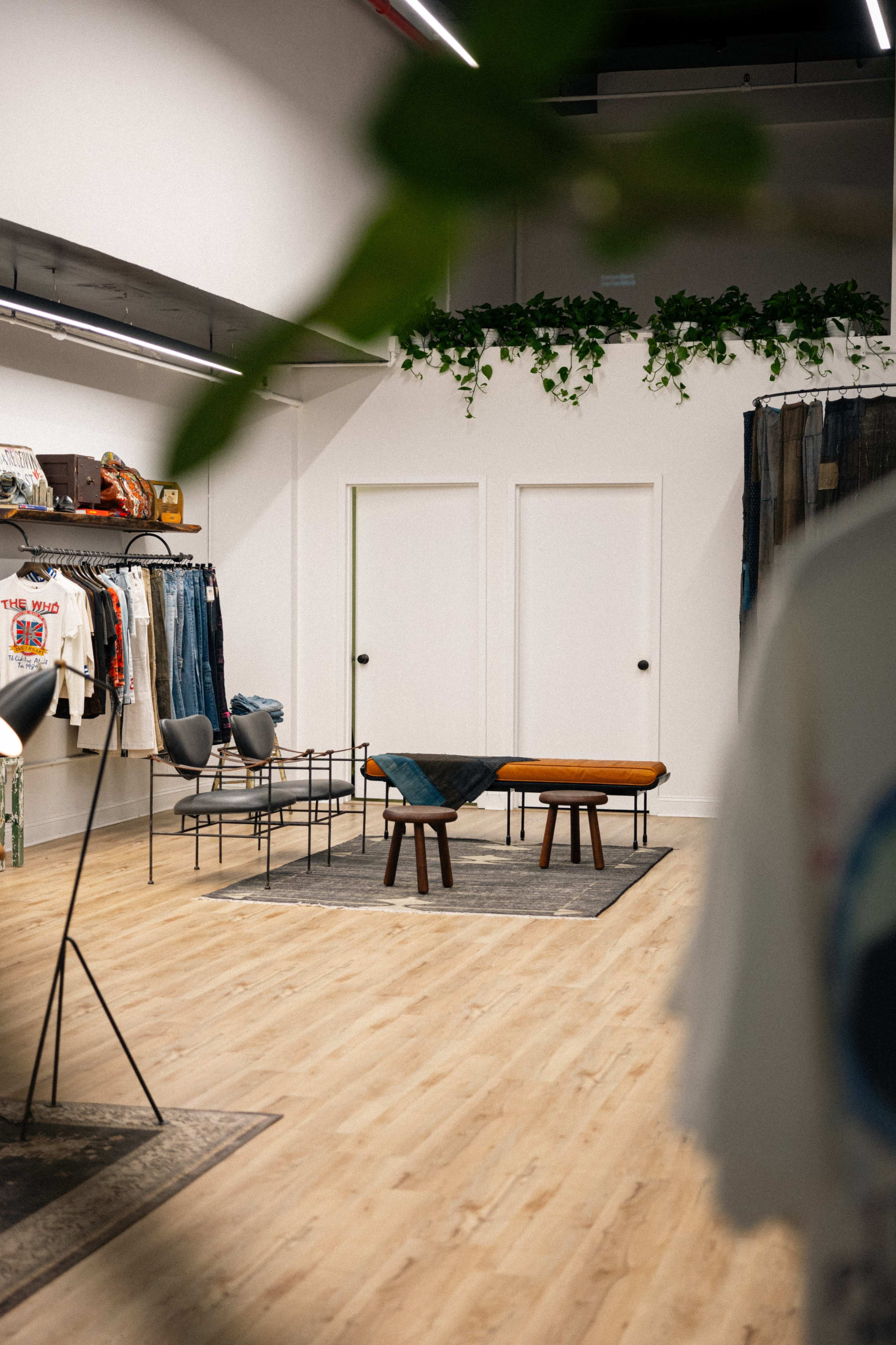 The interior of a clothing store features a seating area with a table and chairs, surrounded by racks of apparel and green plants on the walls.
