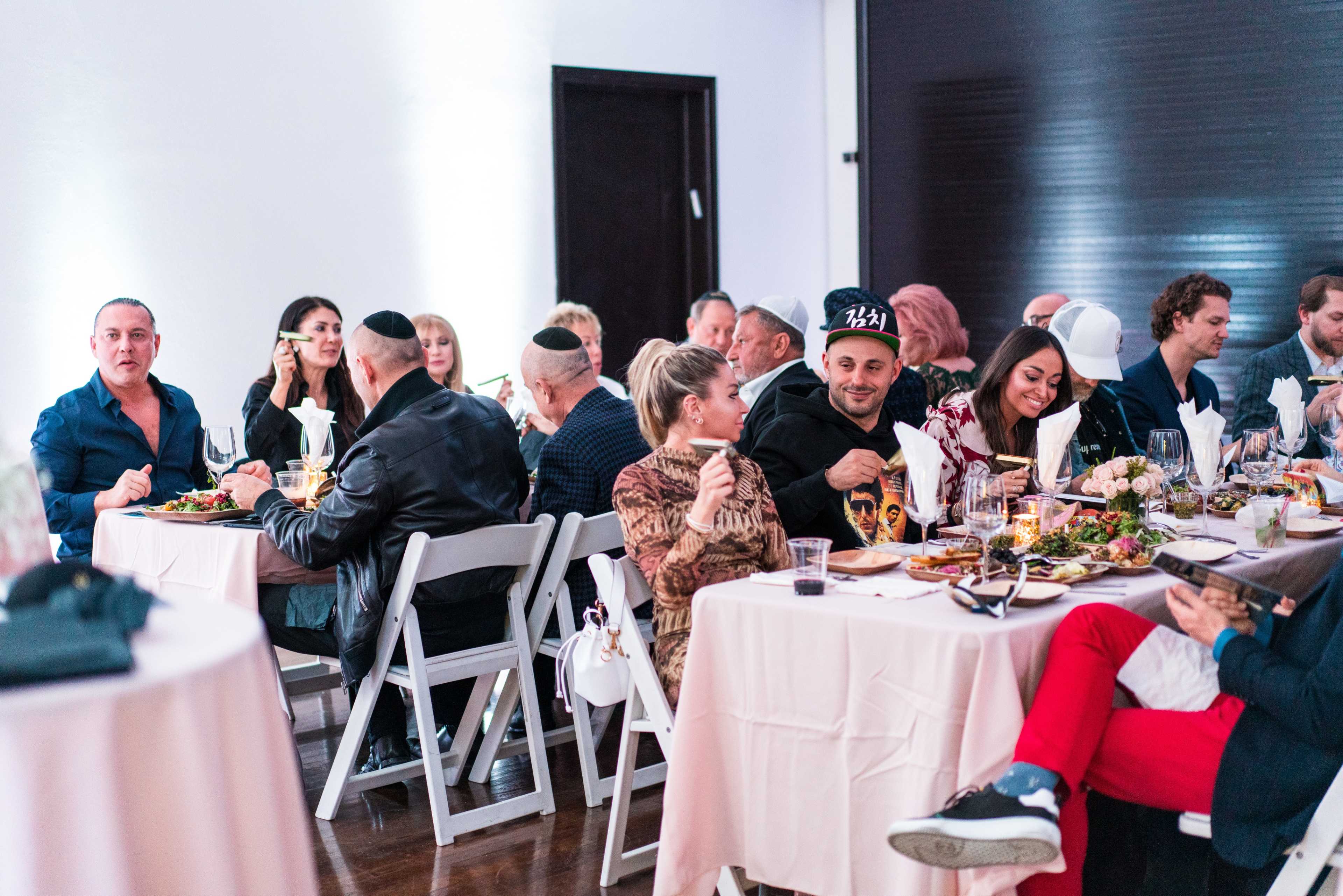 A group of people are seated around tables, enjoying a meal and engaging in conversation at a gathering in a well-lit space.