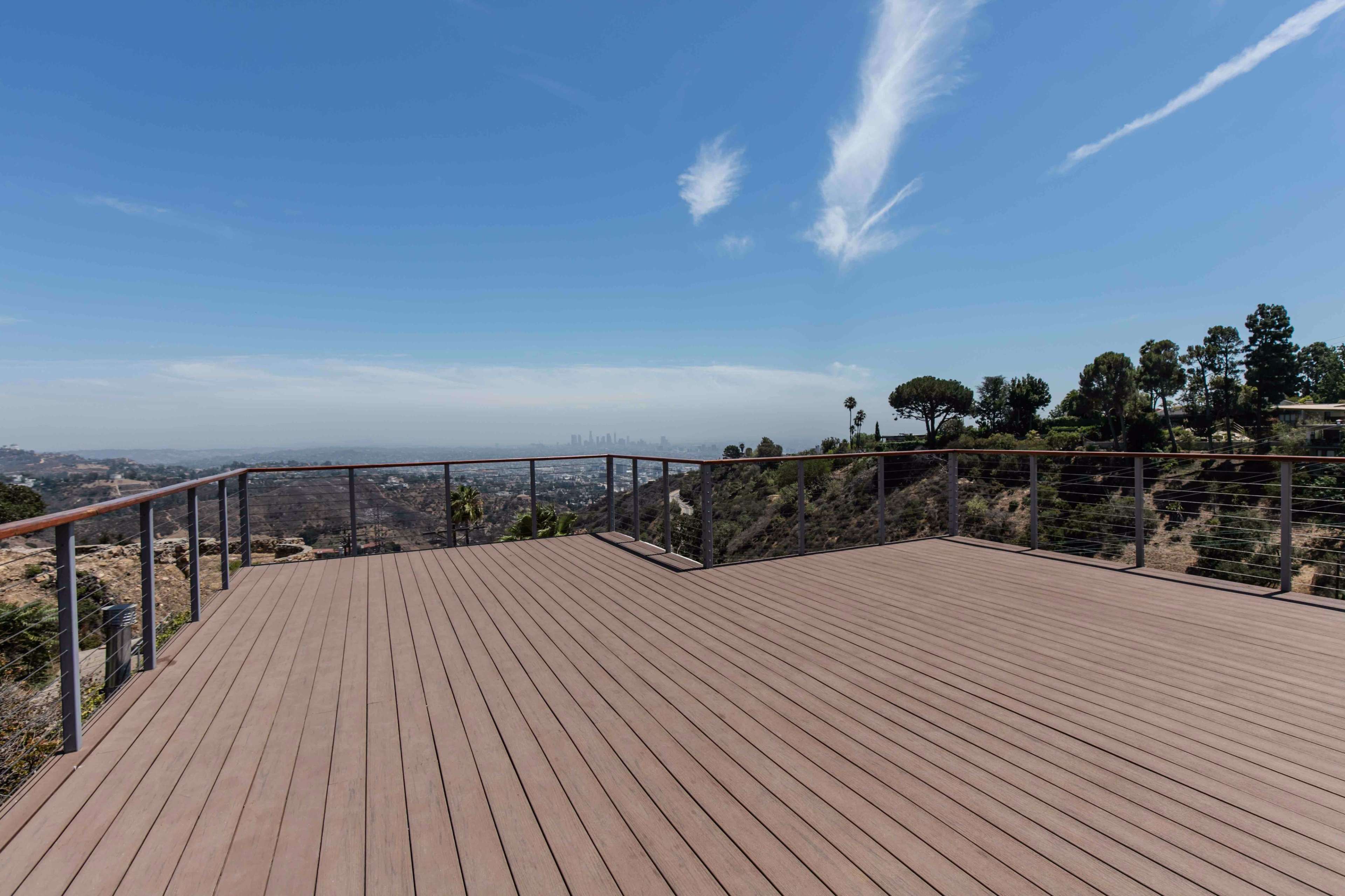 The image shows a spacious wooden deck overlooking a panoramic view of a city and hills under a clear sky.