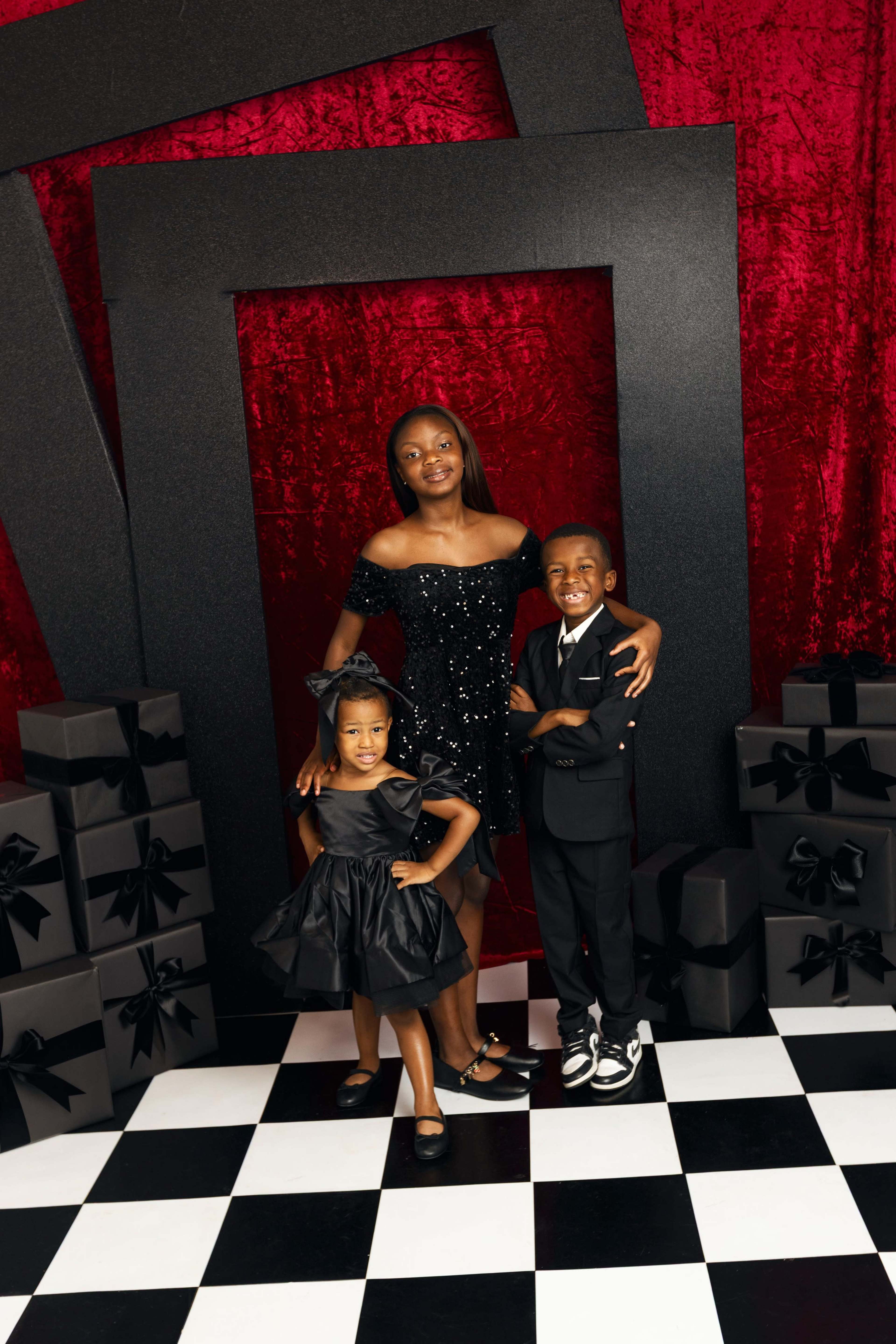 A girl in a black sequin dress stands with two children, one in a formal outfit and the other in a black dress, against a backdrop of red fabric and black gift boxes.