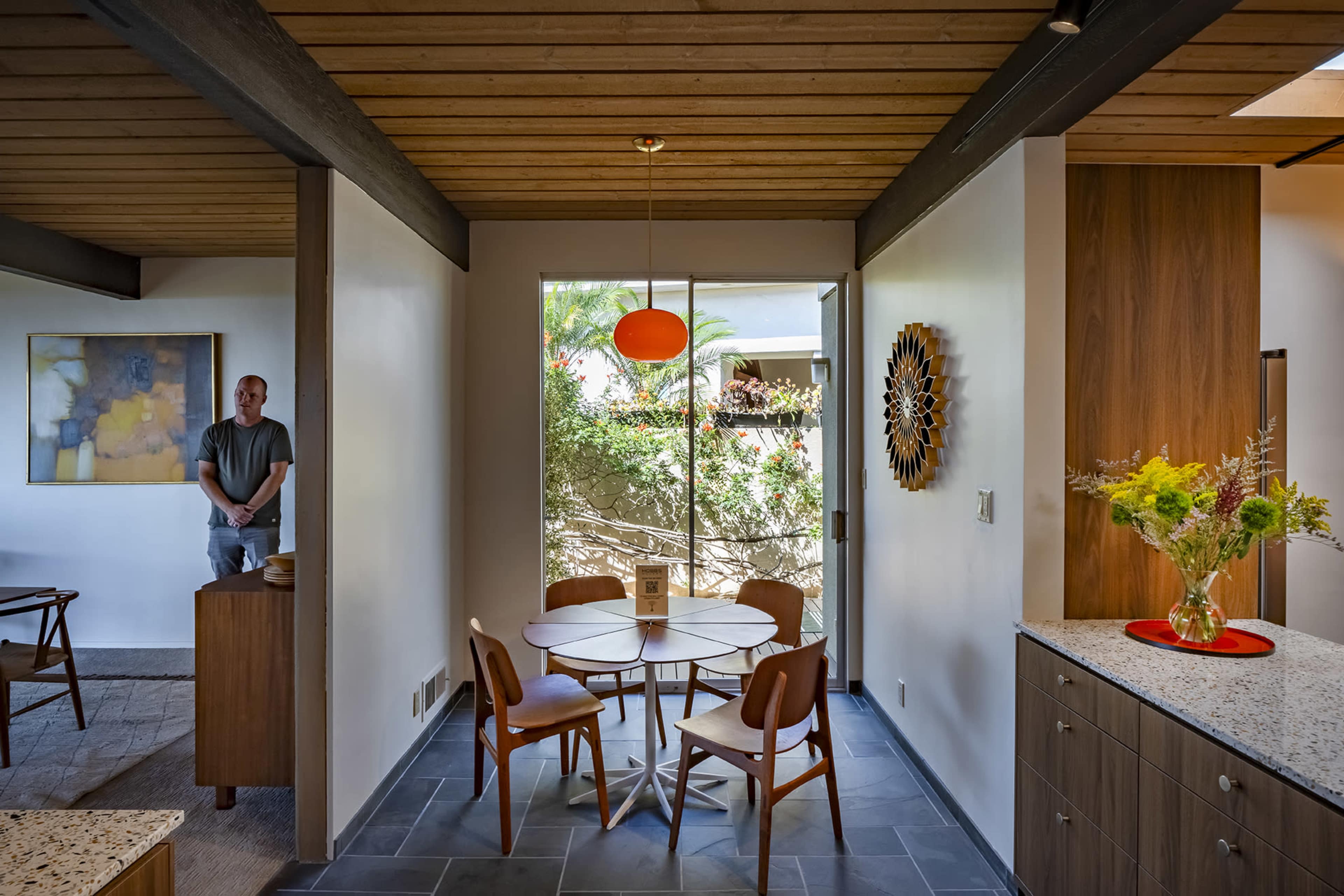 A modern dining area features a circular table with wooden chairs, a colorful pendant light, and a doorway leading to an outdoor garden, while a person stands near a side cabinet.