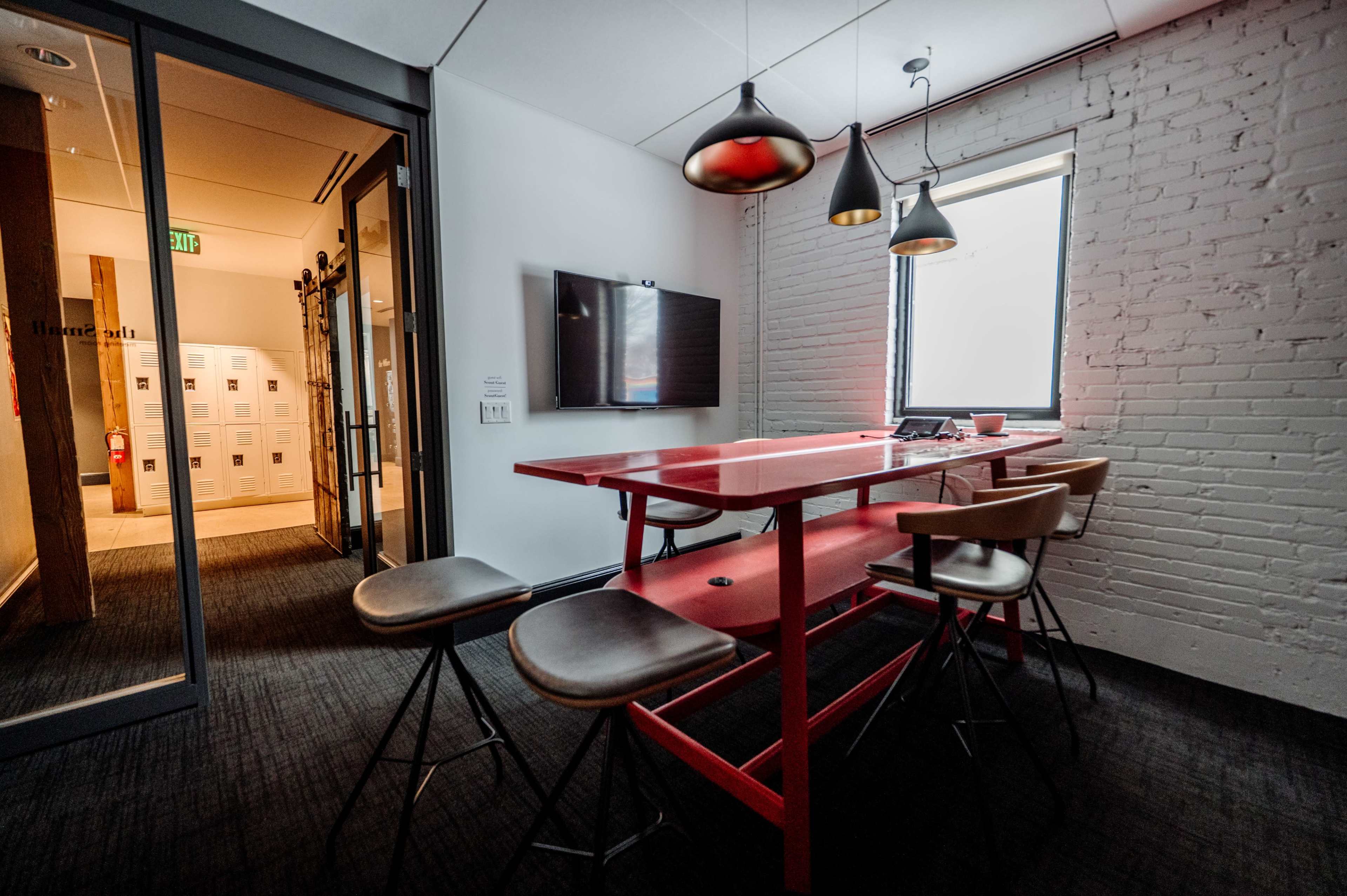 The image shows a modern conference room with a red table, stools, a wall-mounted TV, and a large window providing natural light.
