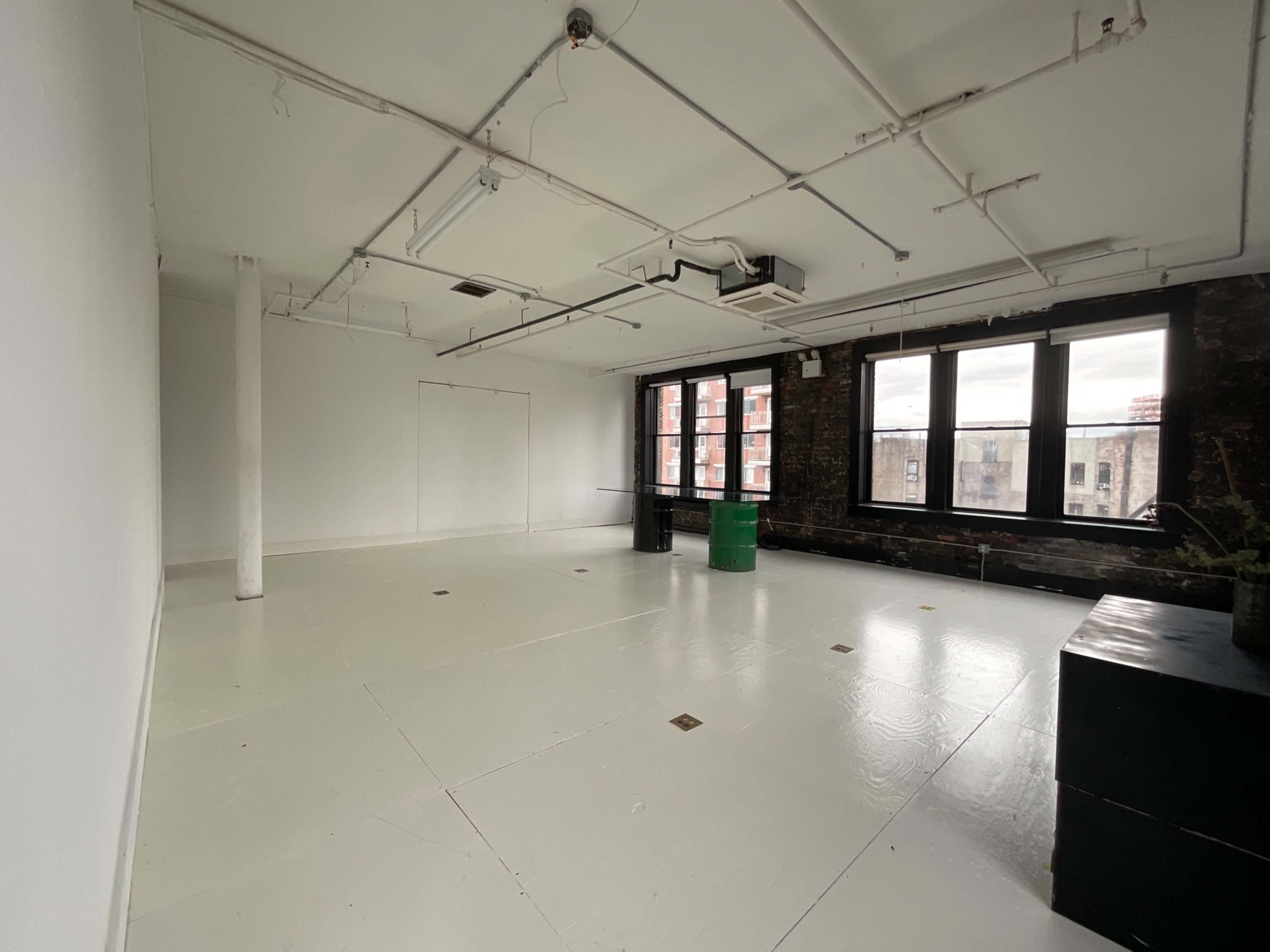 The image shows an empty room with white floors, exposed brick walls, and large windows.