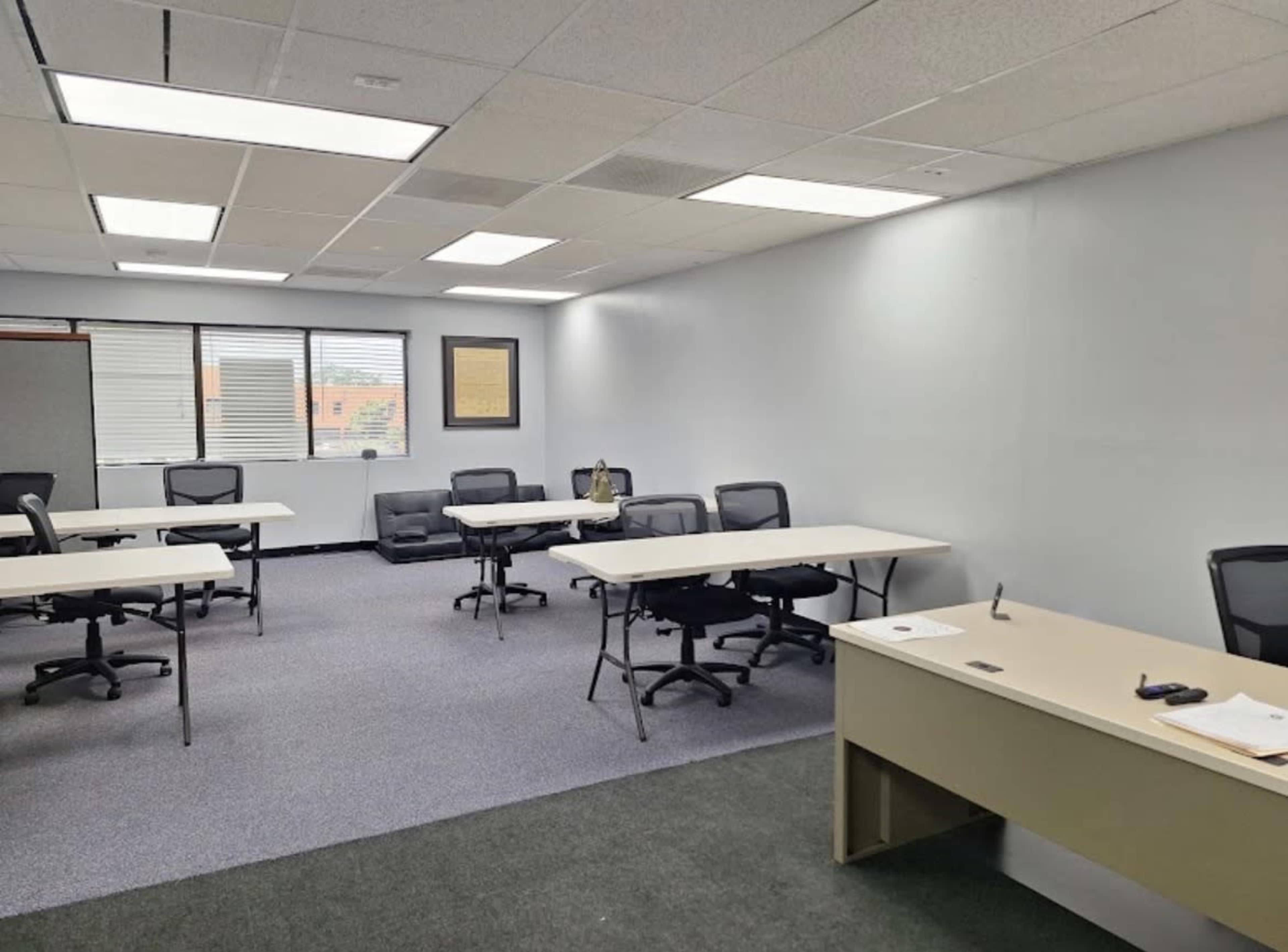 The image shows a spacious, empty classroom or conference room with several tables and chairs arranged in a semi-circle, and a desk is positioned against one wall.