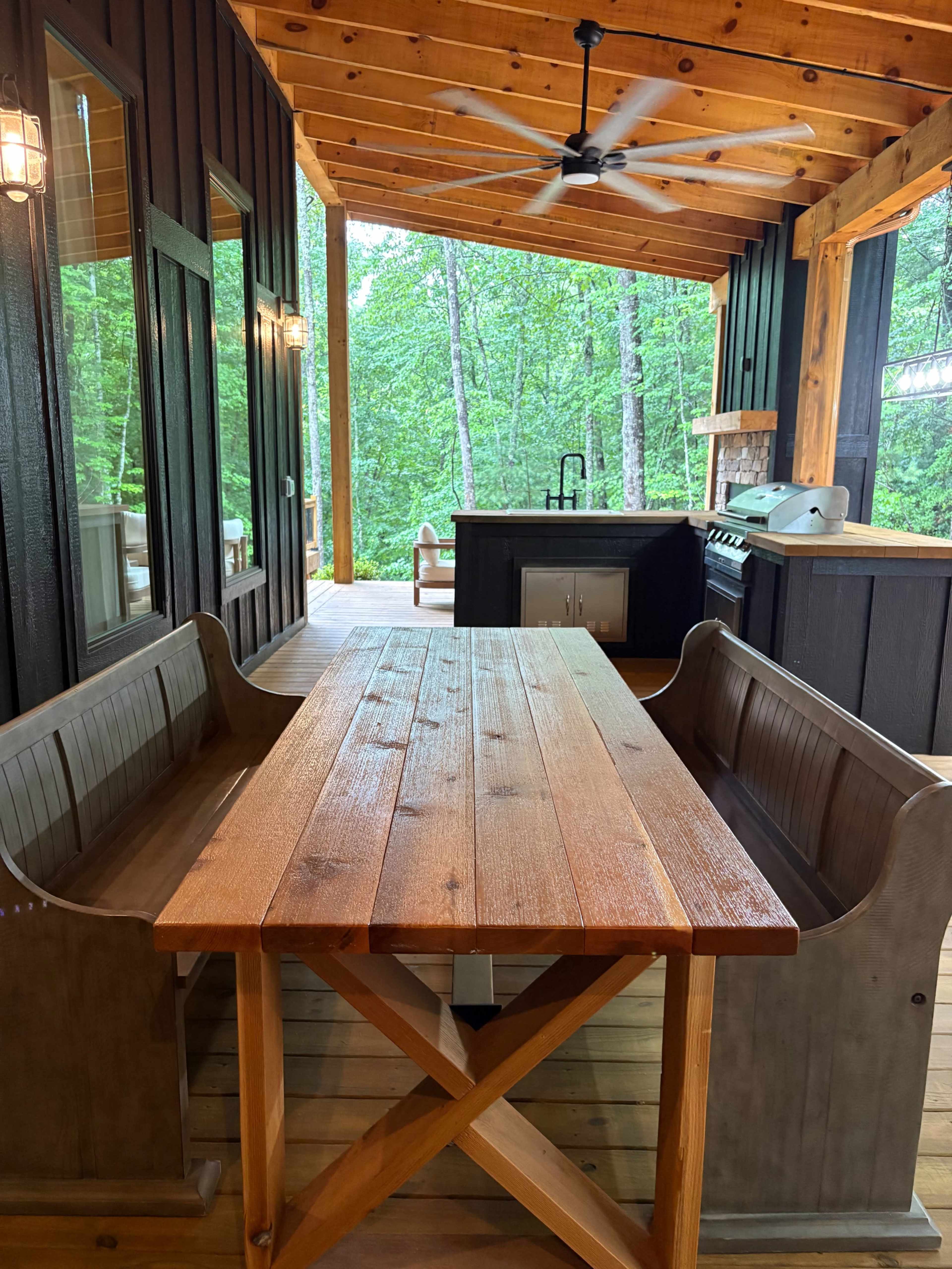 The image shows a wooden dining table surrounded by benches on a covered porch next to a kitchen area, all set in a wooded environment.