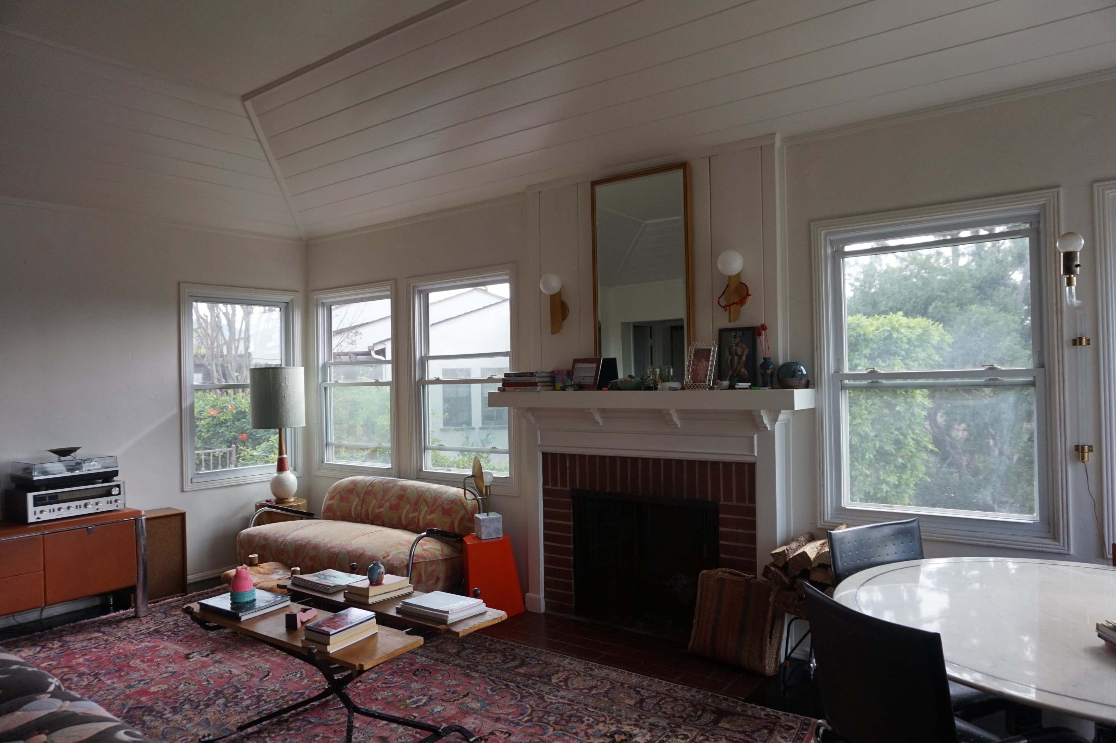 The image shows a living room with a patterned rug, a sofa, a coffee table with books, a fireplace, and large windows providing natural light.