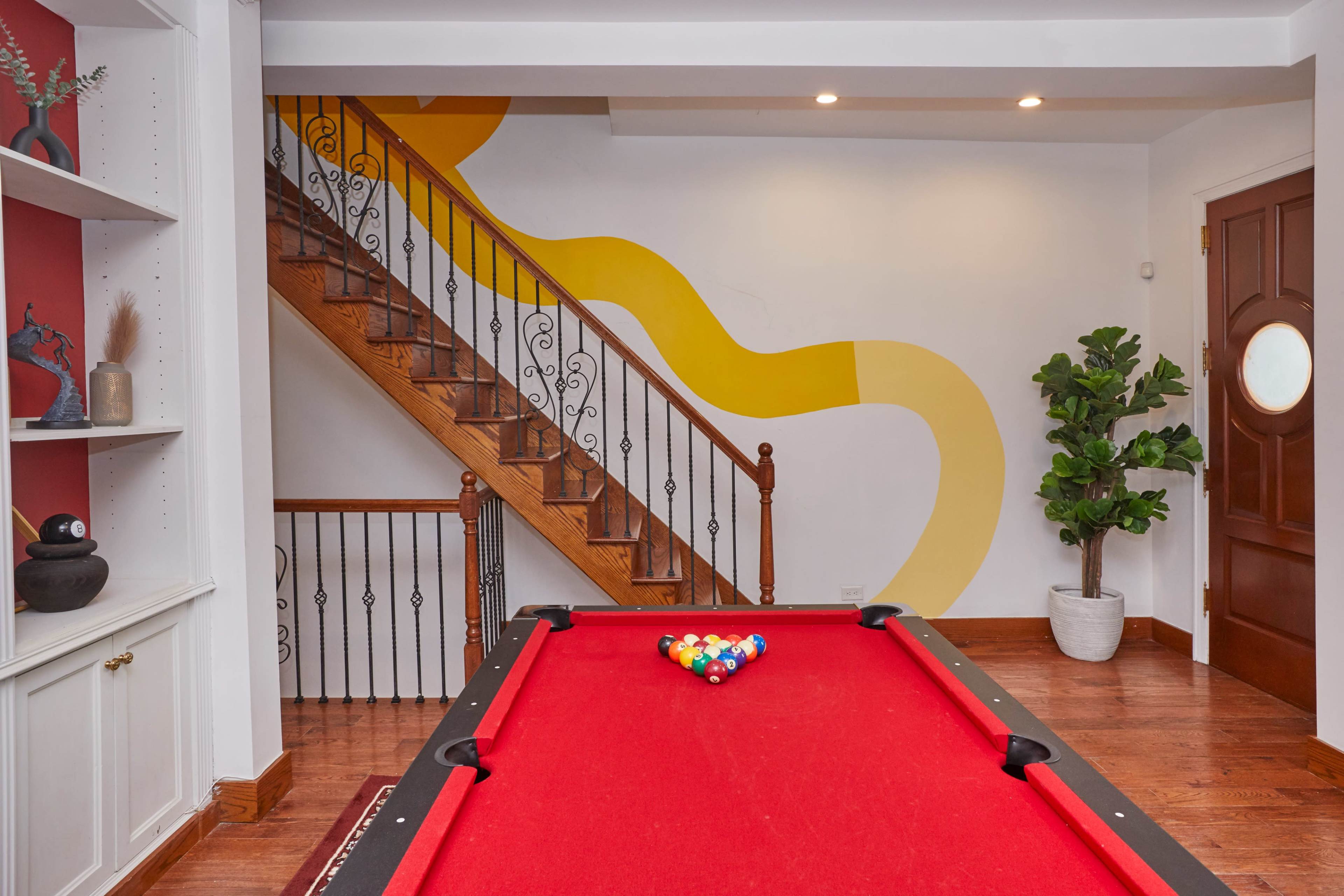 A billiard table with red felt in a room featuring a staircase and a colorful wall design.