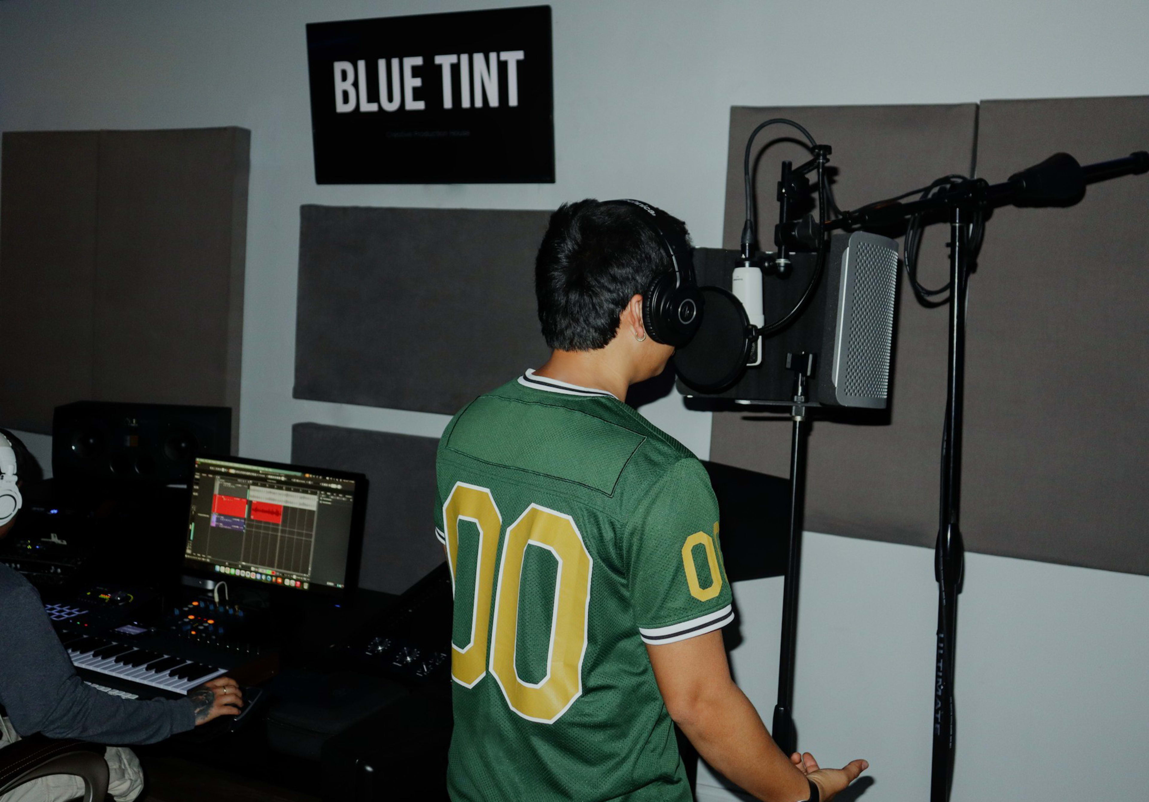 A person wearing a green jersey stands in front of a microphone in a recording studio, while another individual works on music production at a keyboard.
