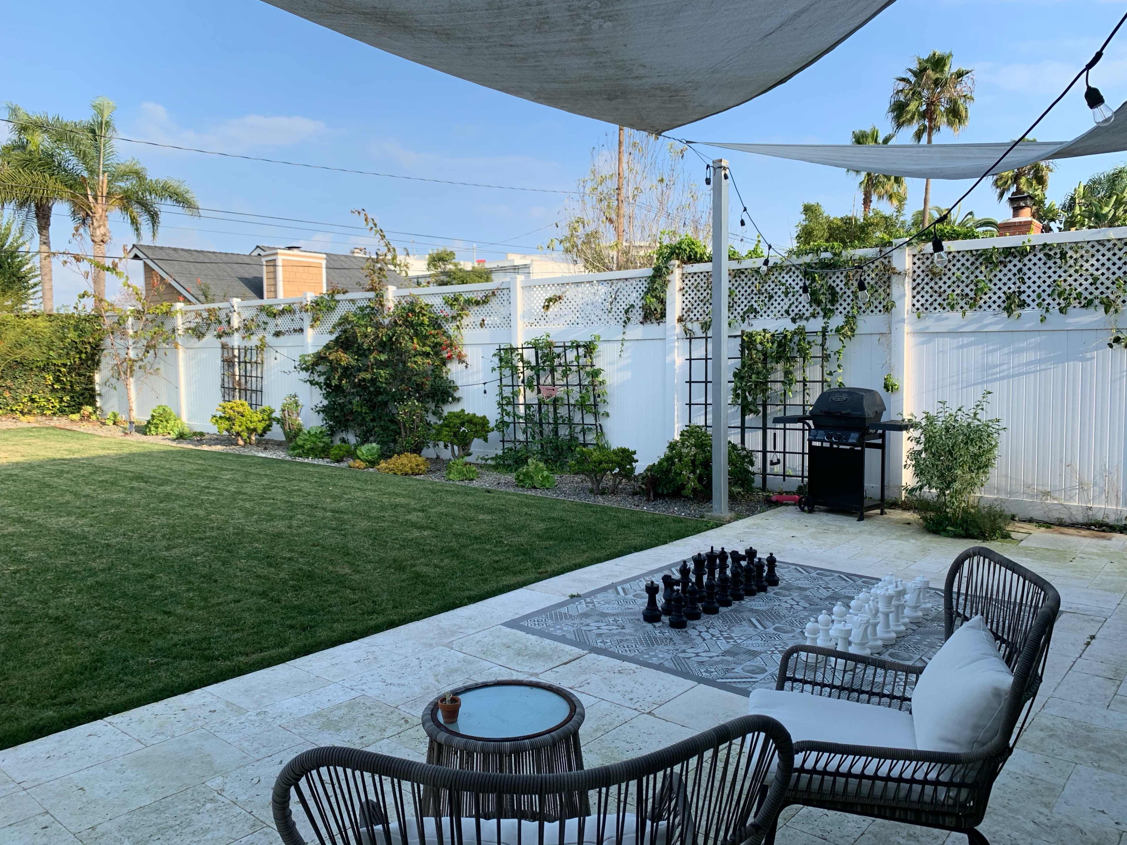 A spacious backyard features a large chess set on a patterned rug, surrounded by green grass, plants, and patio furniture under a shaded area.