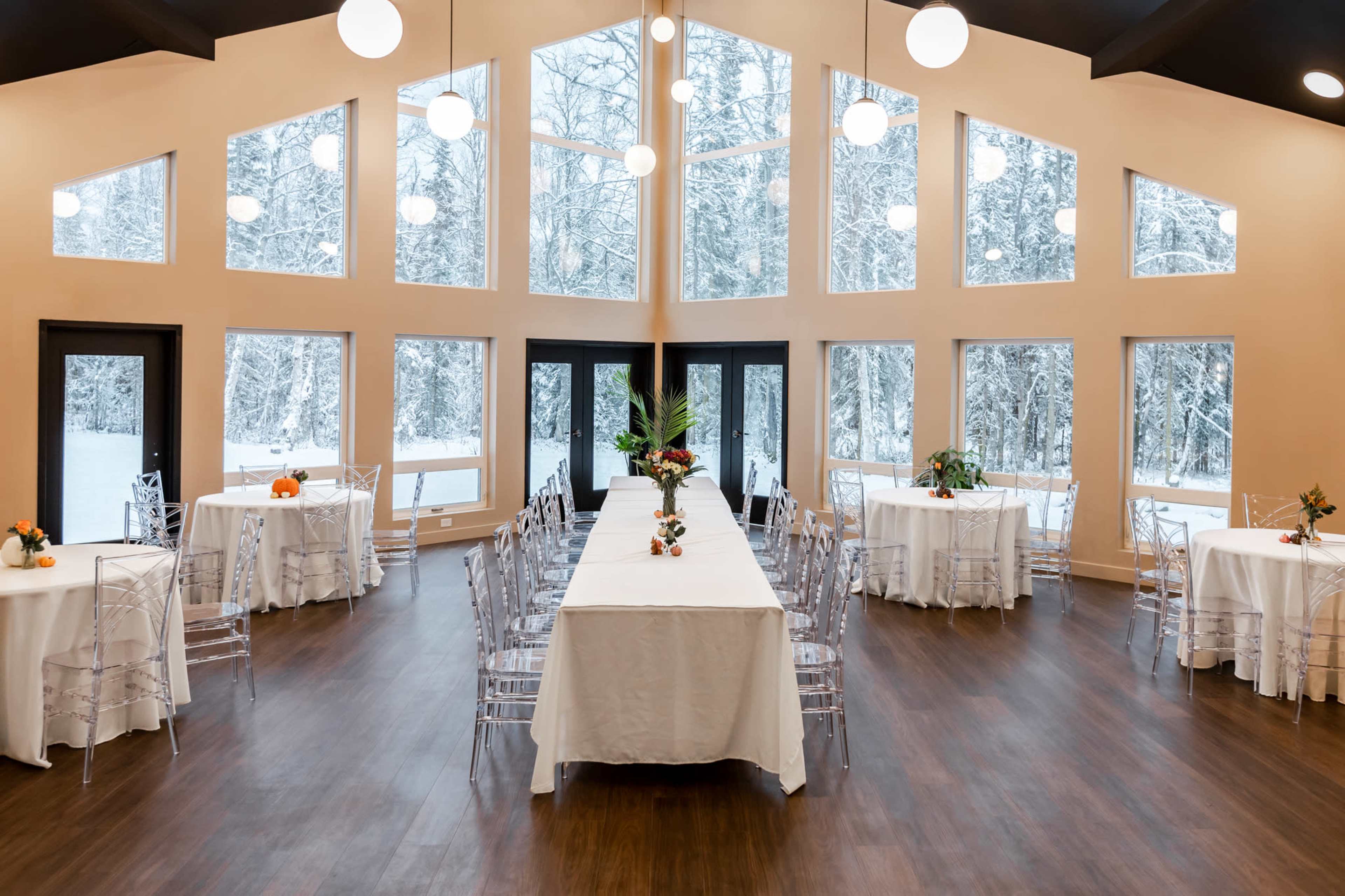 A spacious event venue features several tables dressed with white tablecloths and surrounded by clear acrylic chairs, offering a view of snow-covered trees through large, tall windows.