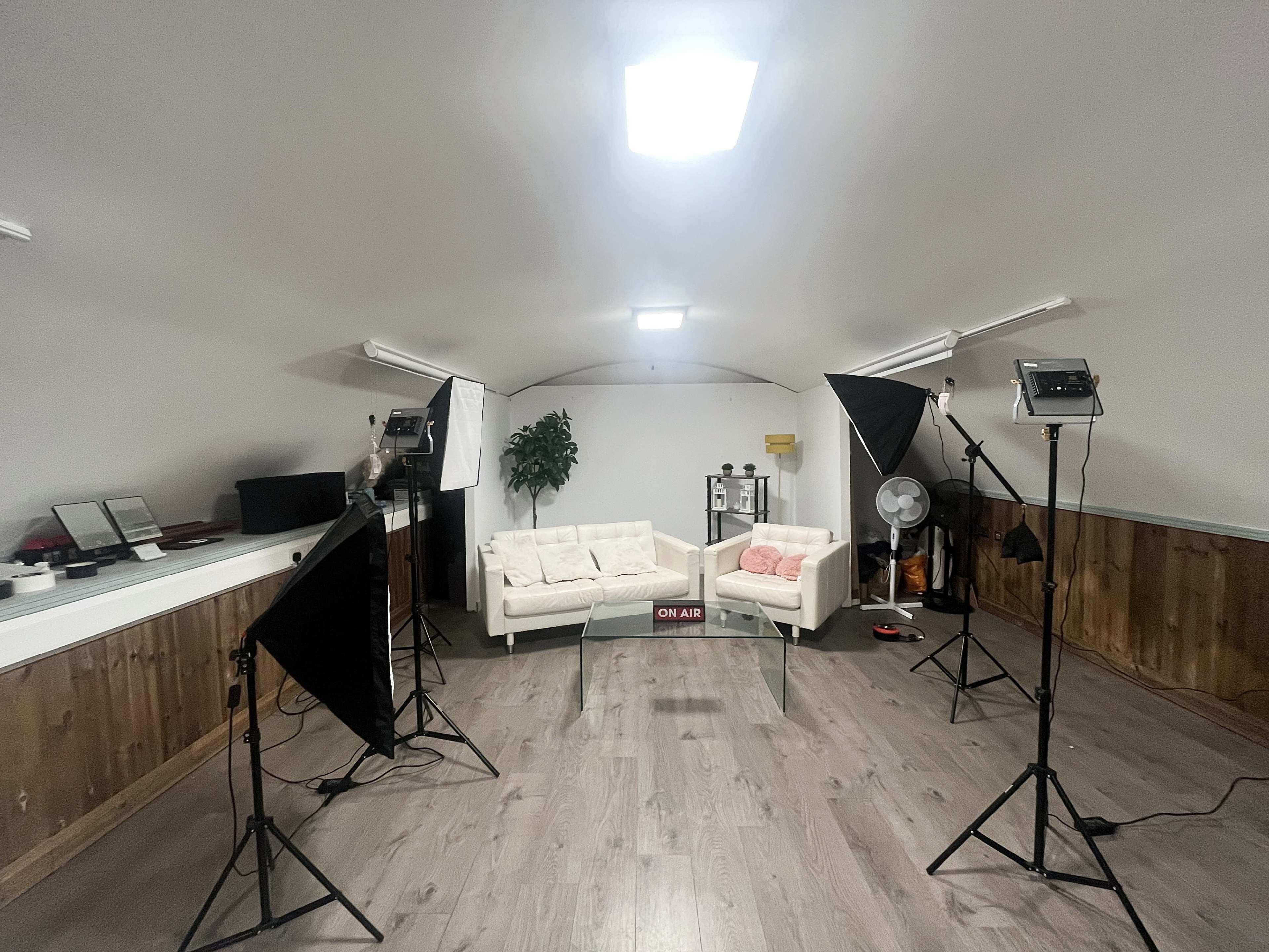 The image shows an indoor studio setup with two white sofas, lighting equipment, and a fan arranged in a spacious room.