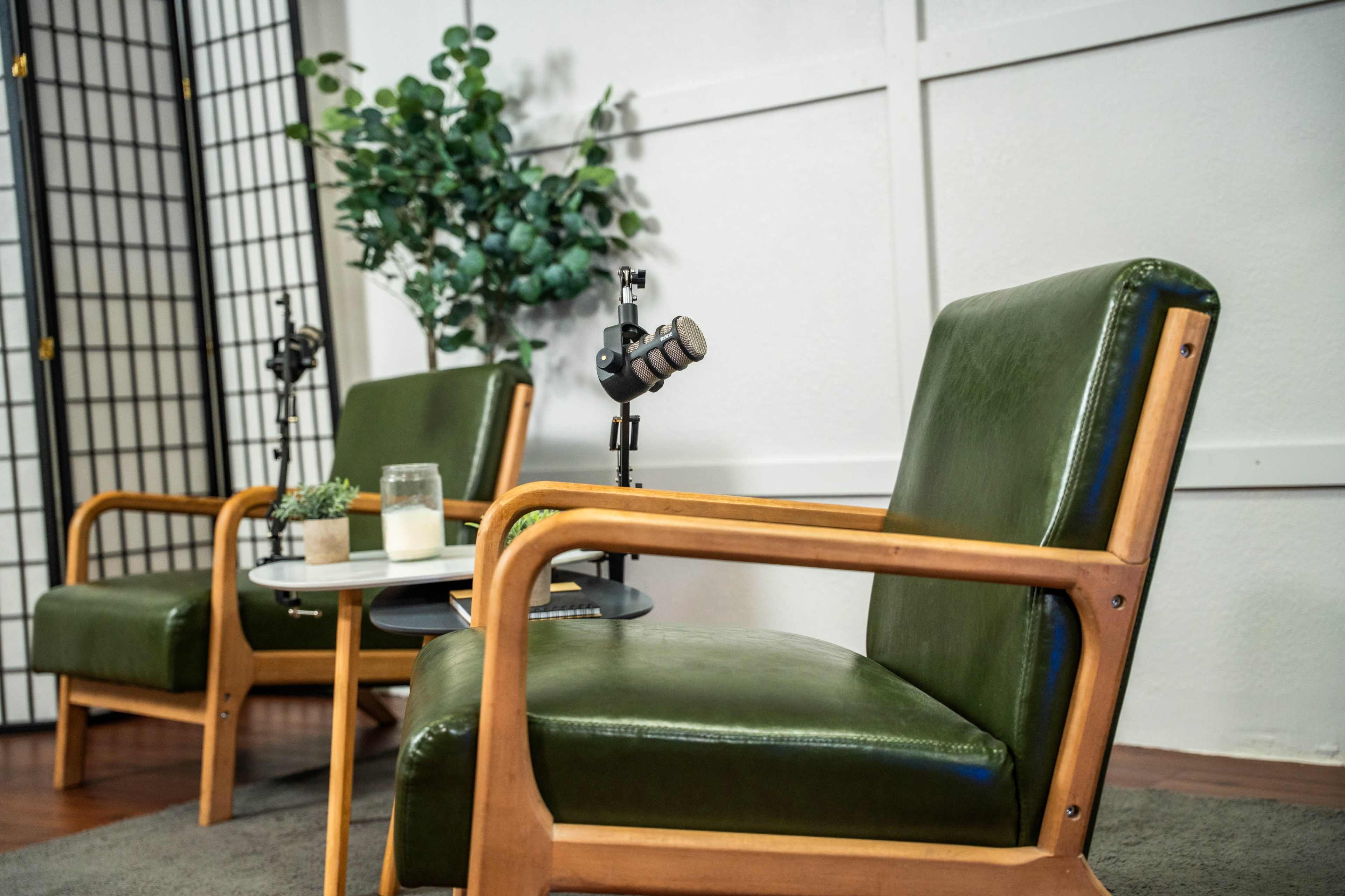 The image shows two green armchairs with wooden frames, a small round table between them, and a plant in the background.