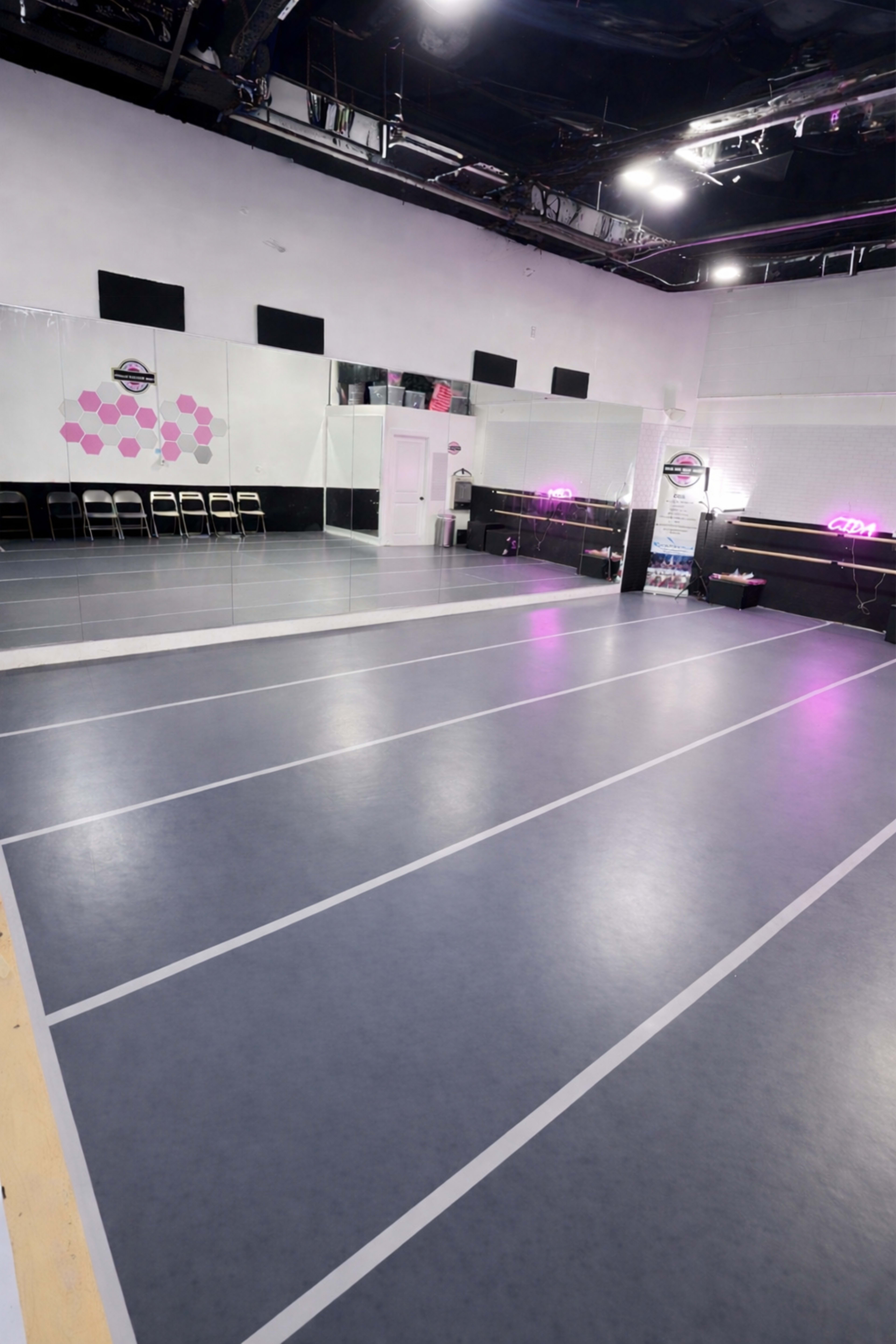 The image shows a spacious dance studio with hardwood flooring, mirrored walls, and a row of folding chairs along one side.