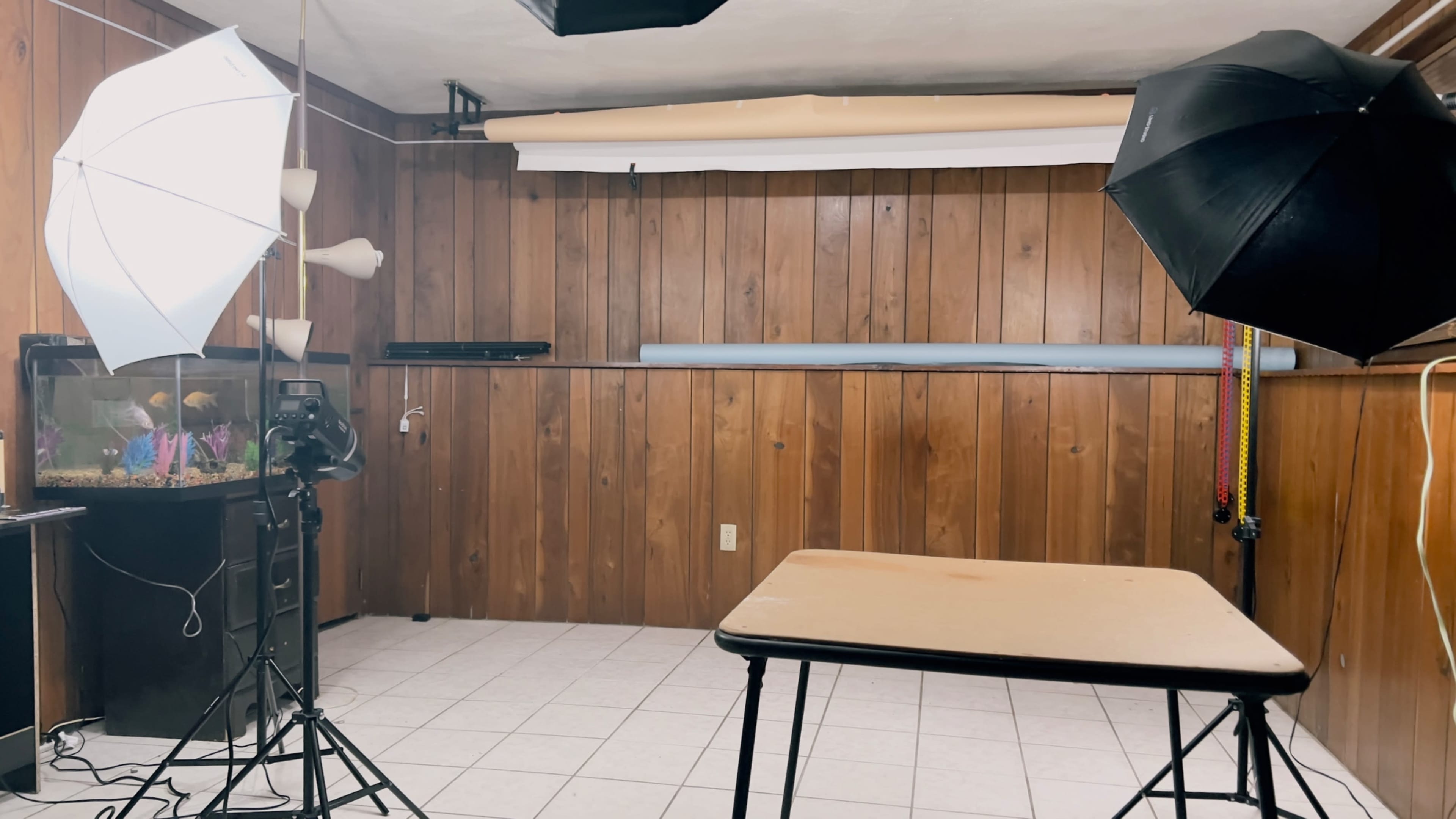 A photography setup in a room with wooden paneling, featuring a table in the center, studio lights on stands, and a fish tank in the corner.