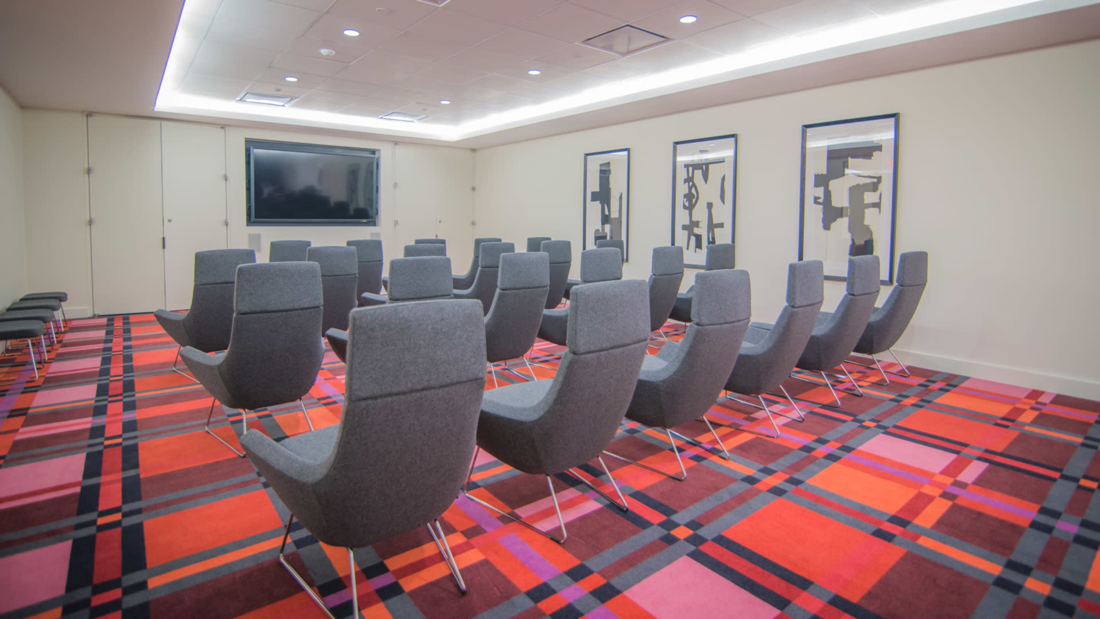 A conference room features a series of gray chairs arranged in rows on a vibrant patterned carpet, with a large screen at the front.
